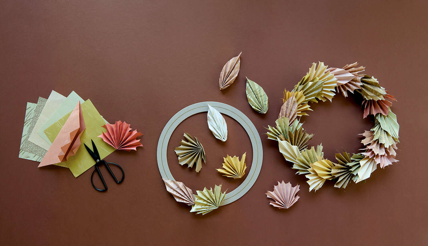 Bastelanleitung Herbstkranz aus Origamipapier zeigt auf braunem Hintergrund Faltpapier, Schere, Kranzrohling und gefaltete Blätter in warmen Pastelltönen
