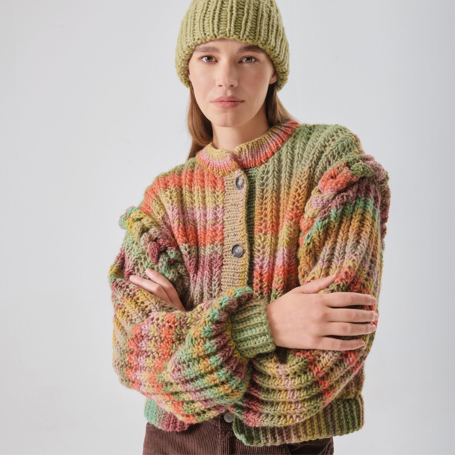 Strickset Mütze Modell 05 aus Made by Me No. 21: Frau trägt bunte Strickjacke und grüne Mütze vor grauem Hintergrund.