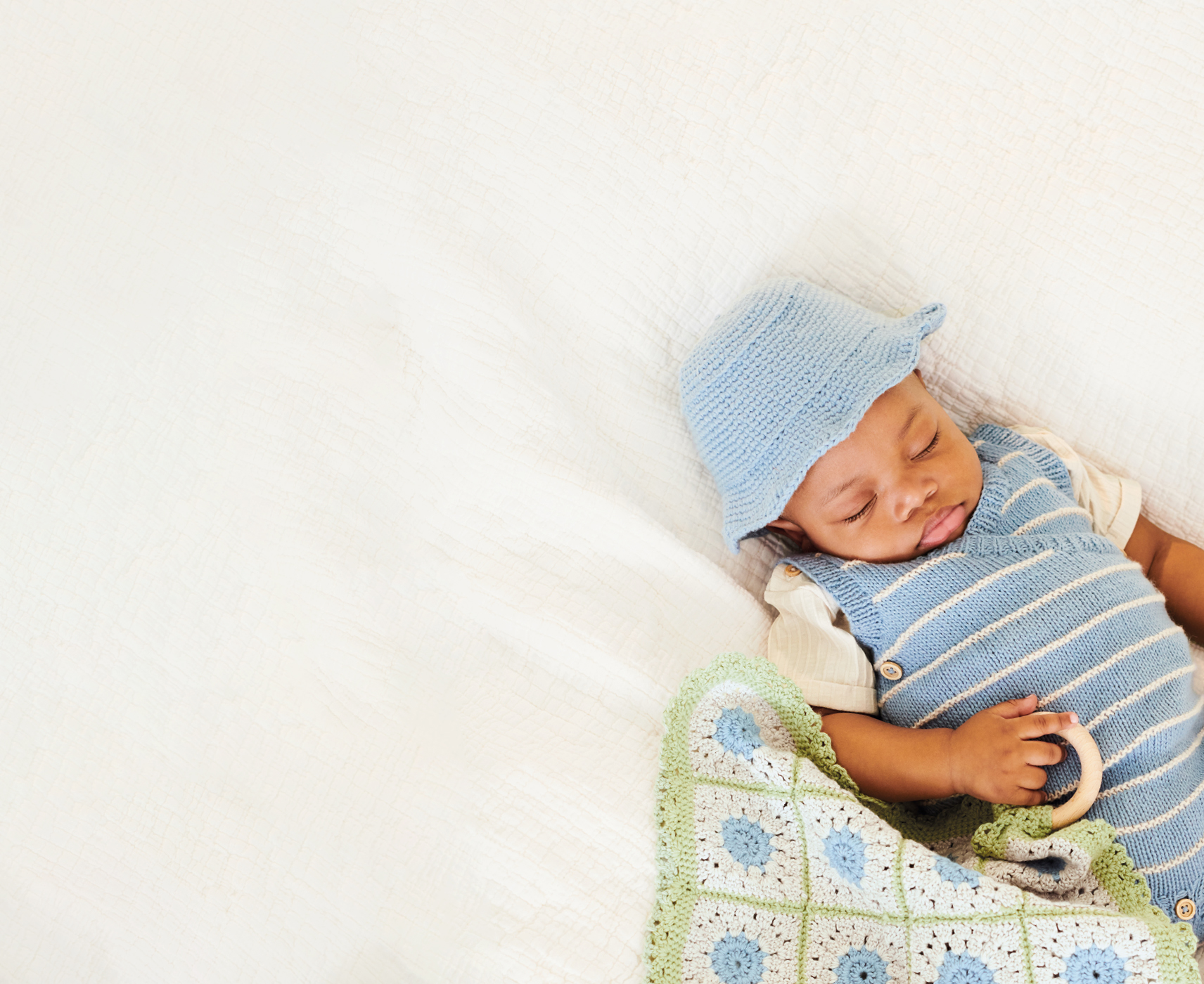 Baby mit gestricktem Outfit und Mütze hält ein gehäkeltes Tuch in der Hand und liegt auf einer Decke.