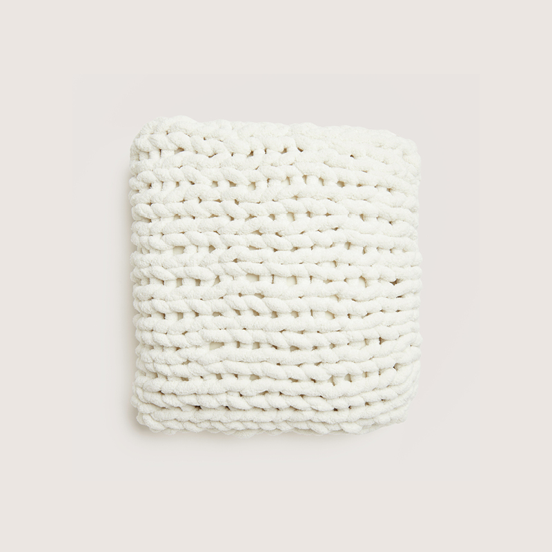 Ein gestricktes Kissen in Weiß liegt vor einem grauen Hintergrund.