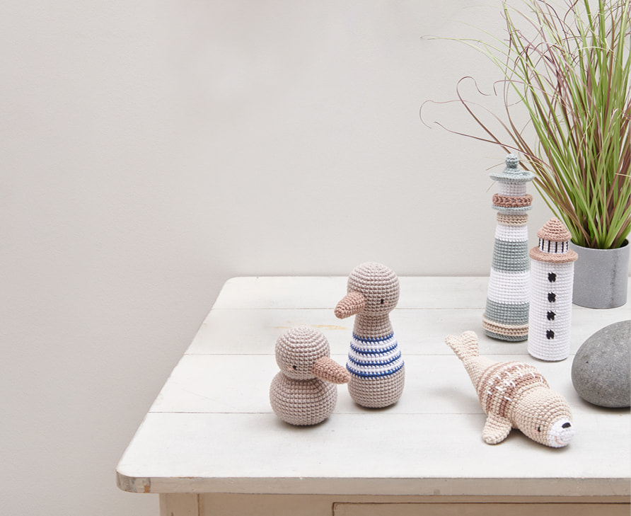 Gehäkelte Figuren mit maritimen Motiven wie Leuchttürme, Möwen und Robbe sind auf einem Tisch angeordnet