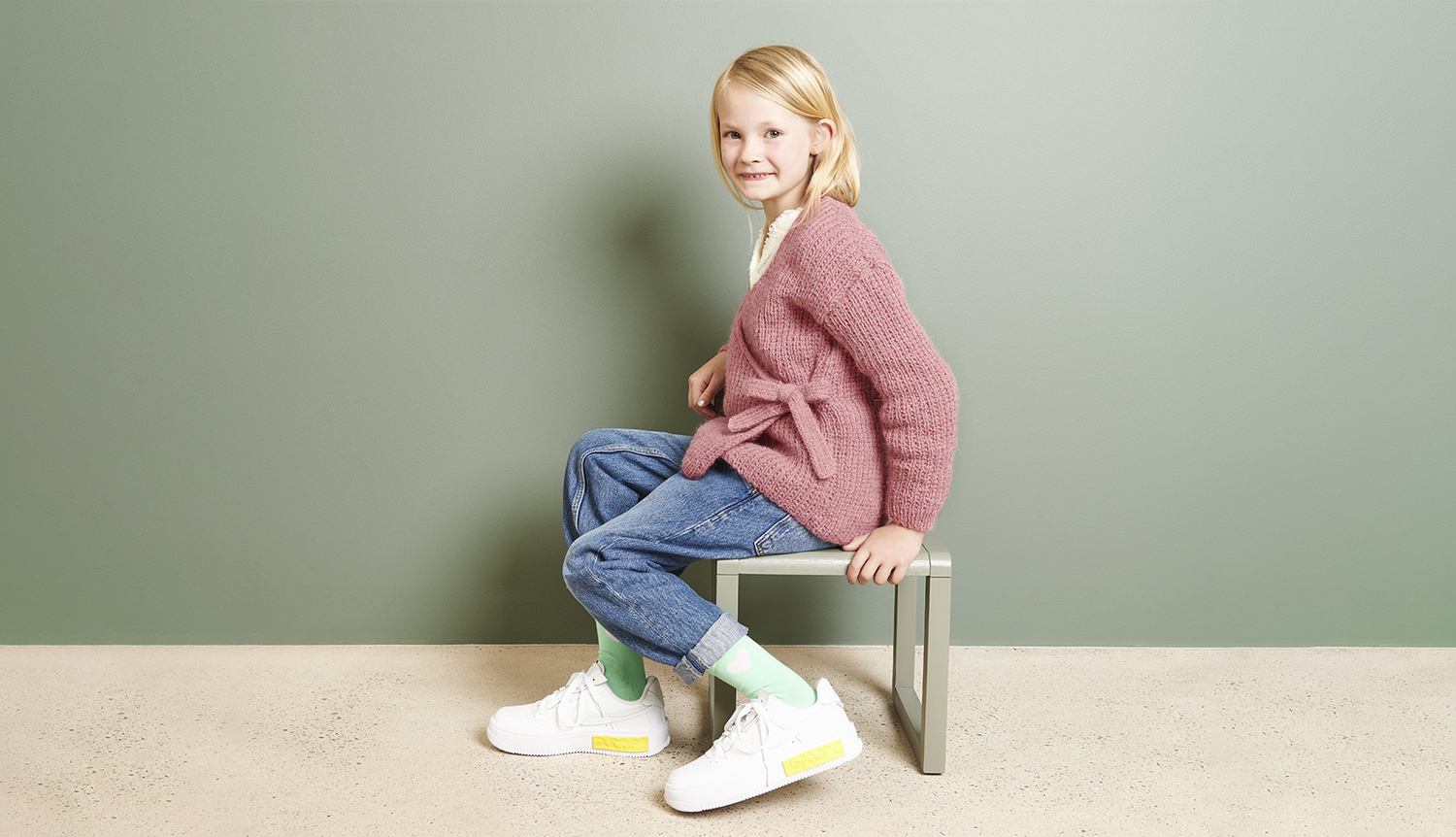Strickanleitung Wickeljacke aus Soft Alpaca: Kind in rosa Jacke, Jeans und weißen Sneakers sitzt auf einer Bank vor grünem Hintergrund.
