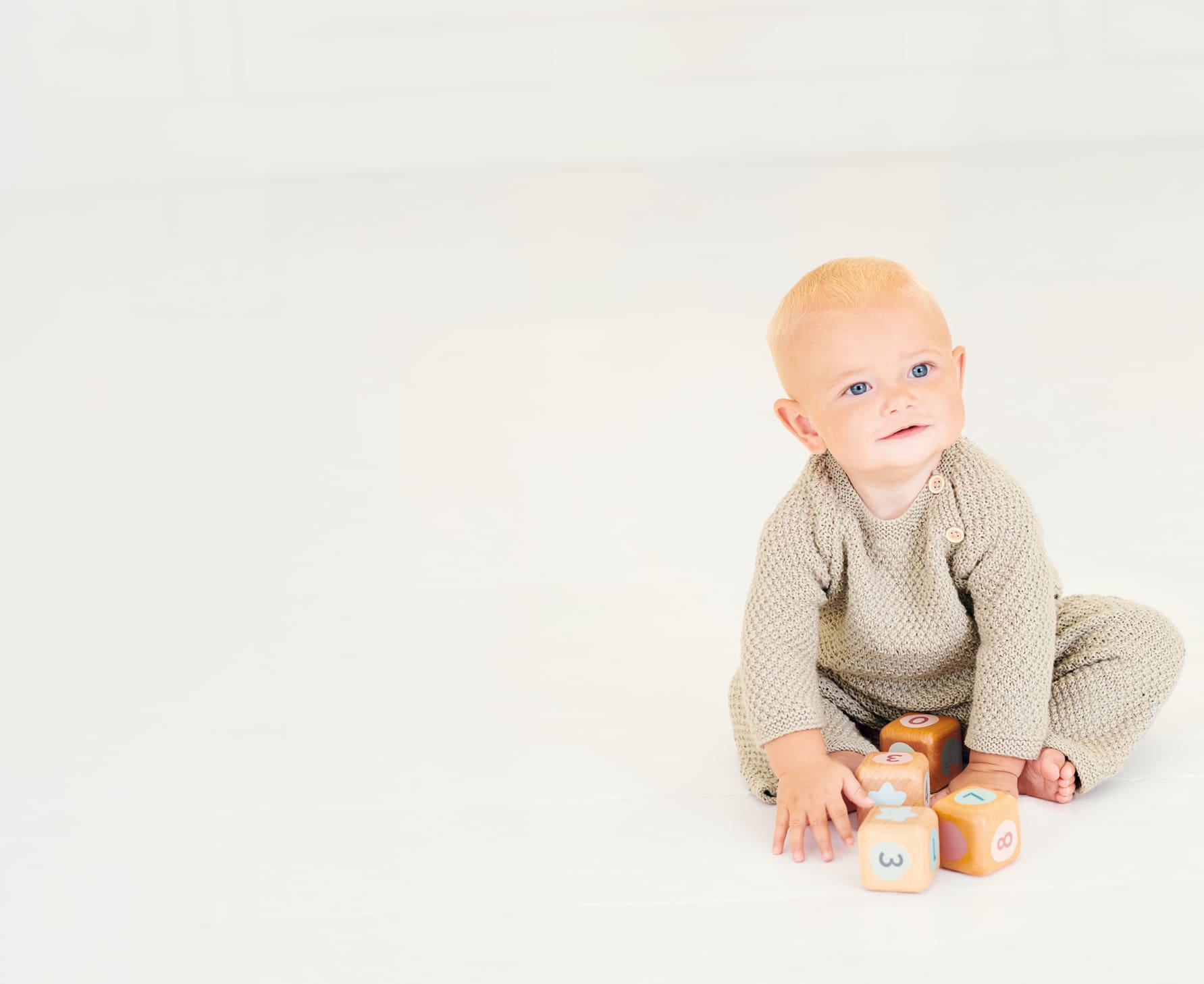 Ein Baby im gestrickten Outfit sitzt mit Spielzeugwürfeln auf dem Boden.