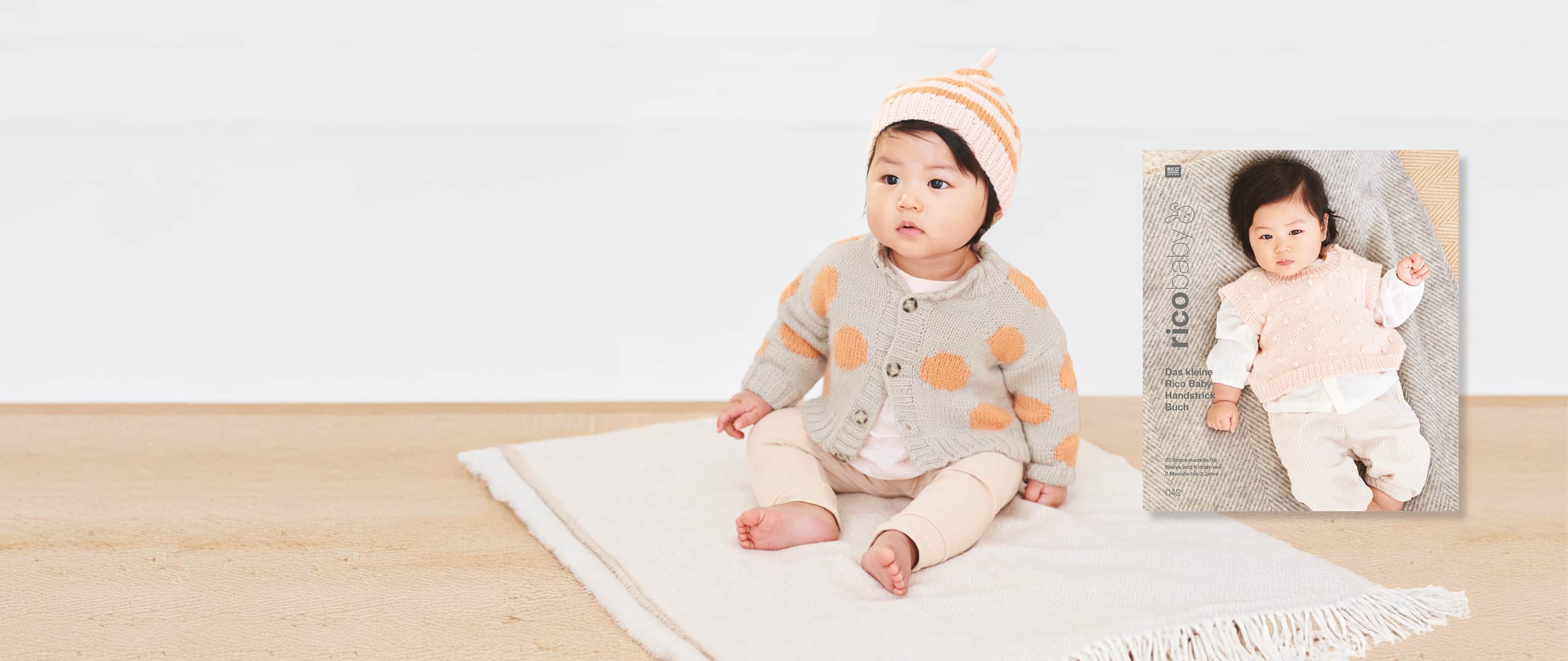 Ein Baby in gestrickter Jacke mit Punkten und gestreifter Mütze sitzt auf einer Decke