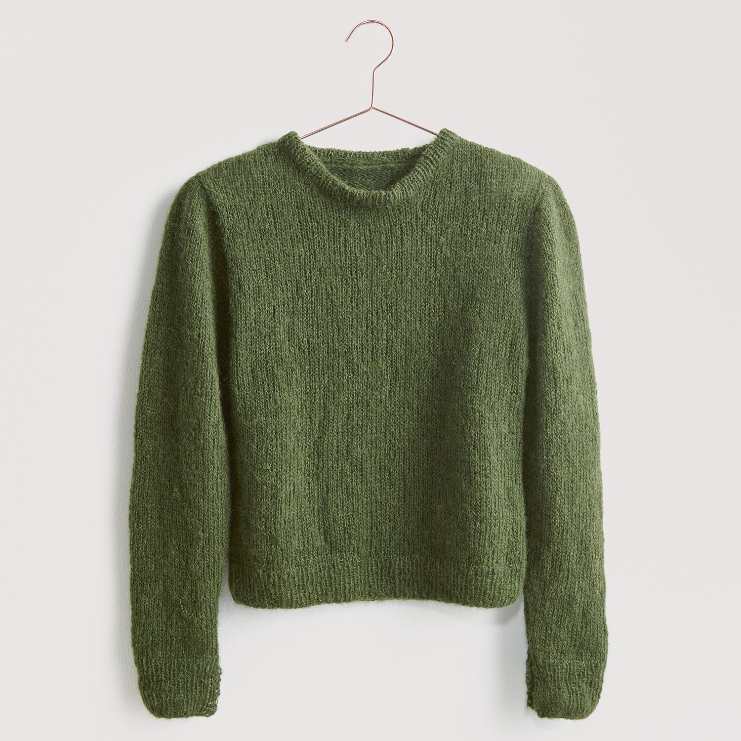 Luxury Knits - Premium Natural Fibres Sprachauswahl deutsch von Rico Design Strickset Pullover Modell 21 aus Luxury Knits: Grüner Pullover aus natürlichen Fasern, hängend an einem Kleiderbügel vor grauem Hintergrund.