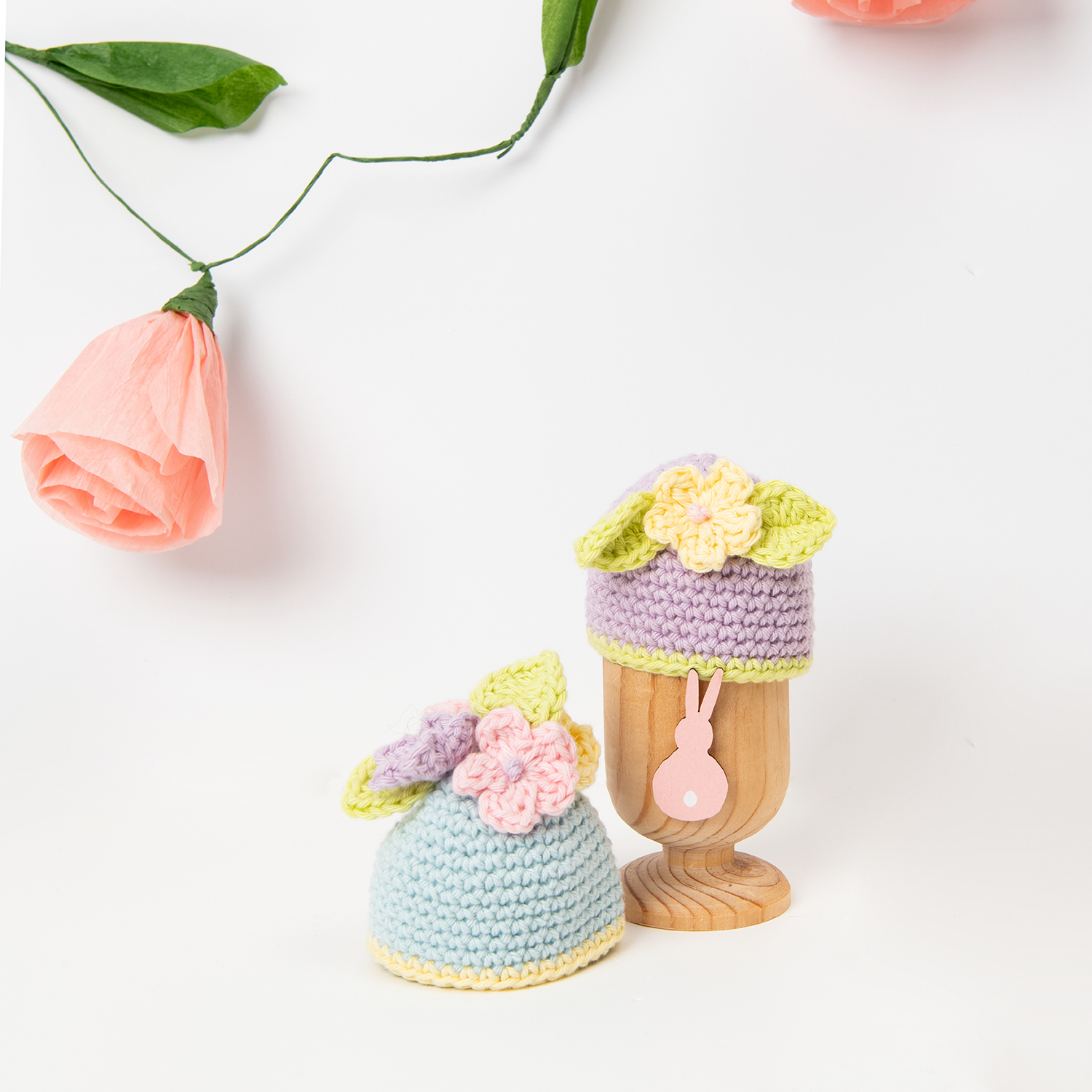 Häkelanleitung Ricorumi Eierwärmer: Zwei pastellfarbene Eierwärmer mit Blumenmotiv auf Holzständer.
