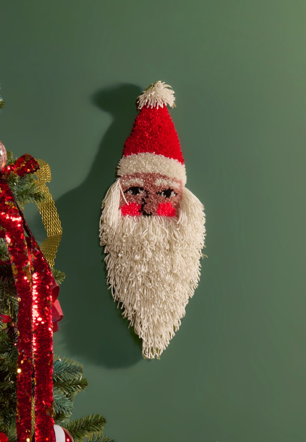 Ein geknüpfter Wandbehang in Form eines Weihnachtsmanns hängt an einer dunkelgrünen Wand. 