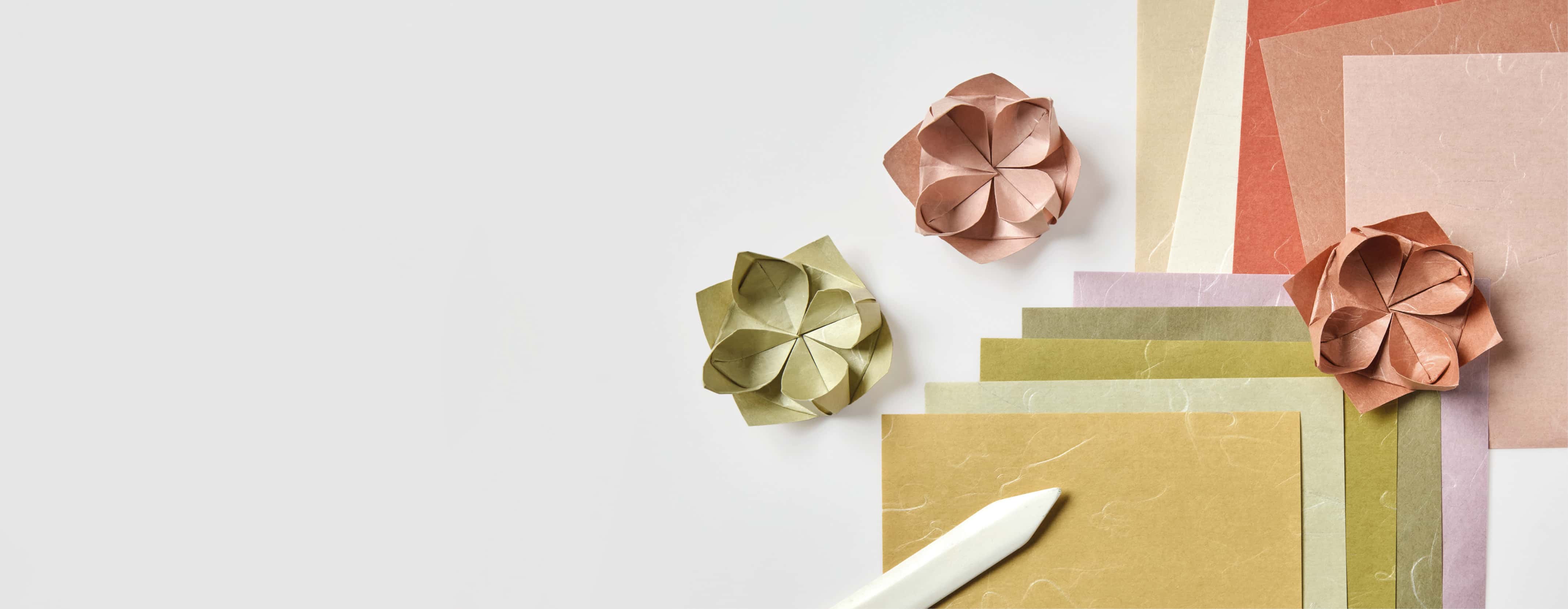 Origamipapier in erdigen Tönen und gefaltete Papierblumen liegen auf grauem Hintergrund. 