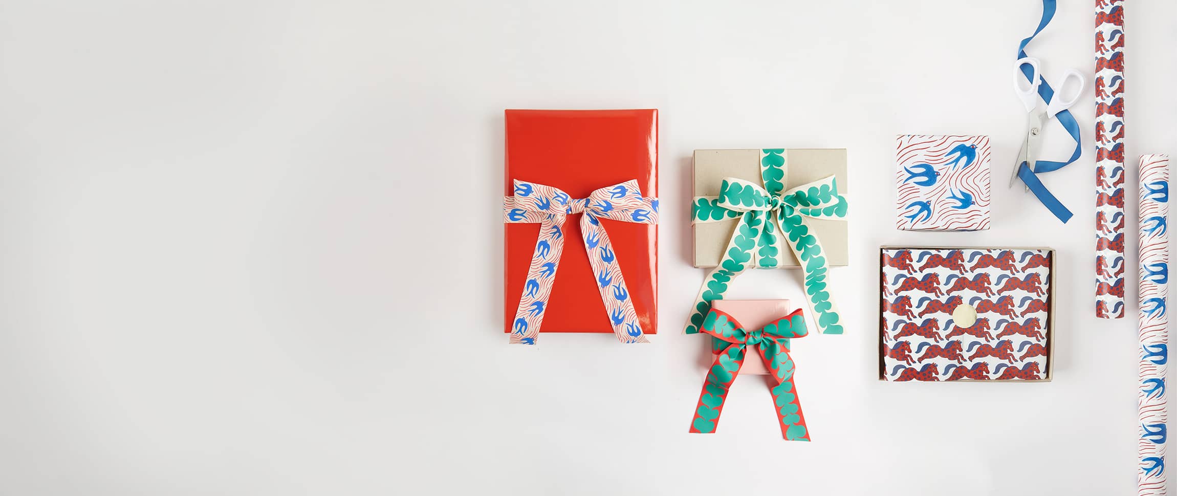 Verpackte Geschenke und Geschenkpapierrollen mit Designs aus der Kollektion Midsommar Magic liegen auf grauem Hintergrund