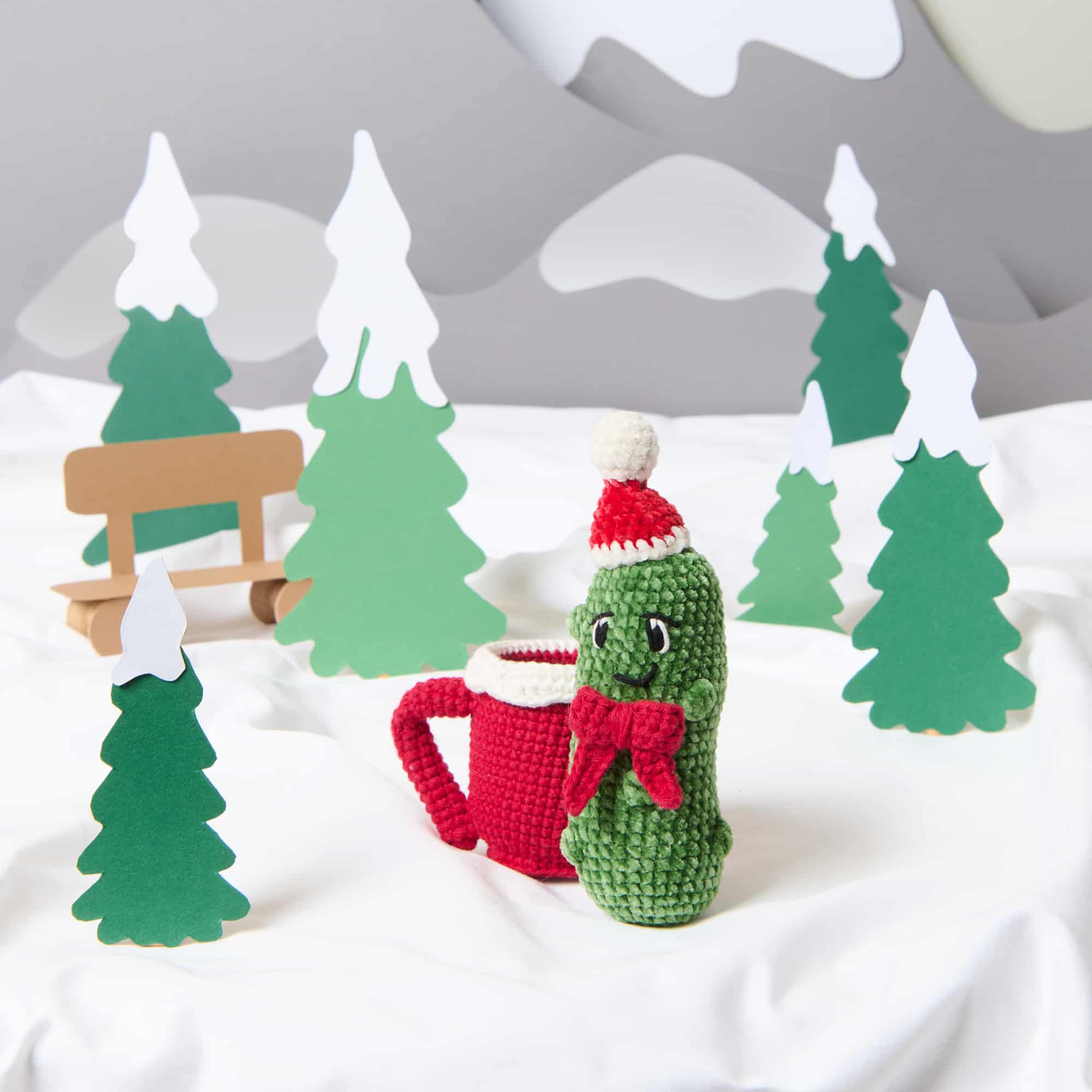 Gehäkelte Gurke mit Weihnachtsmütze und eine gehäkelte Tasse stehen in einer Winter-Landschaft.