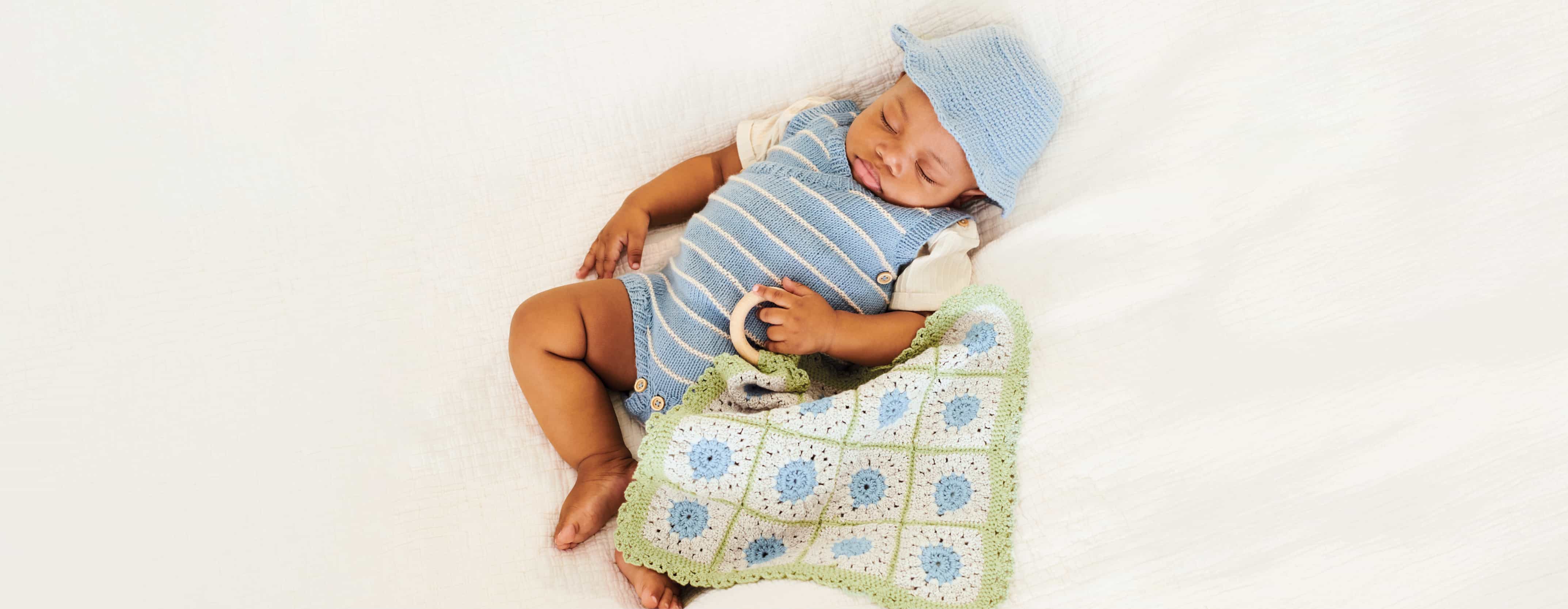 Baby mit gestricktem Outfit und Mütze hält ein gehäkeltes Tuch in der Hand und liegt auf einer Decke.