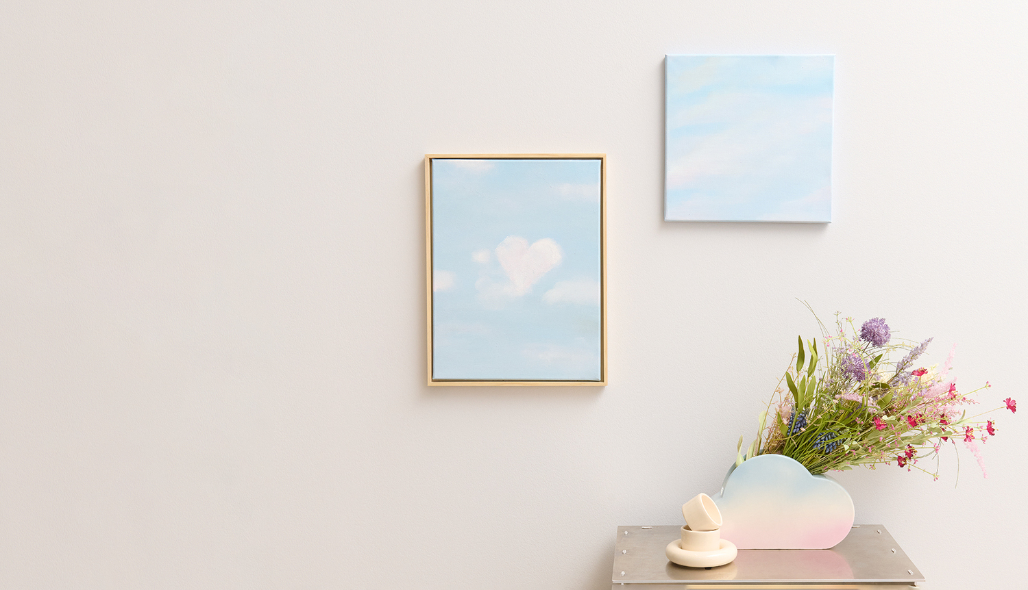 Anleitung Wolkenbild mit Acrylfarbe malen: Zwei blaue Gemälde an weißer Wand, daneben Vase mit Blumen.