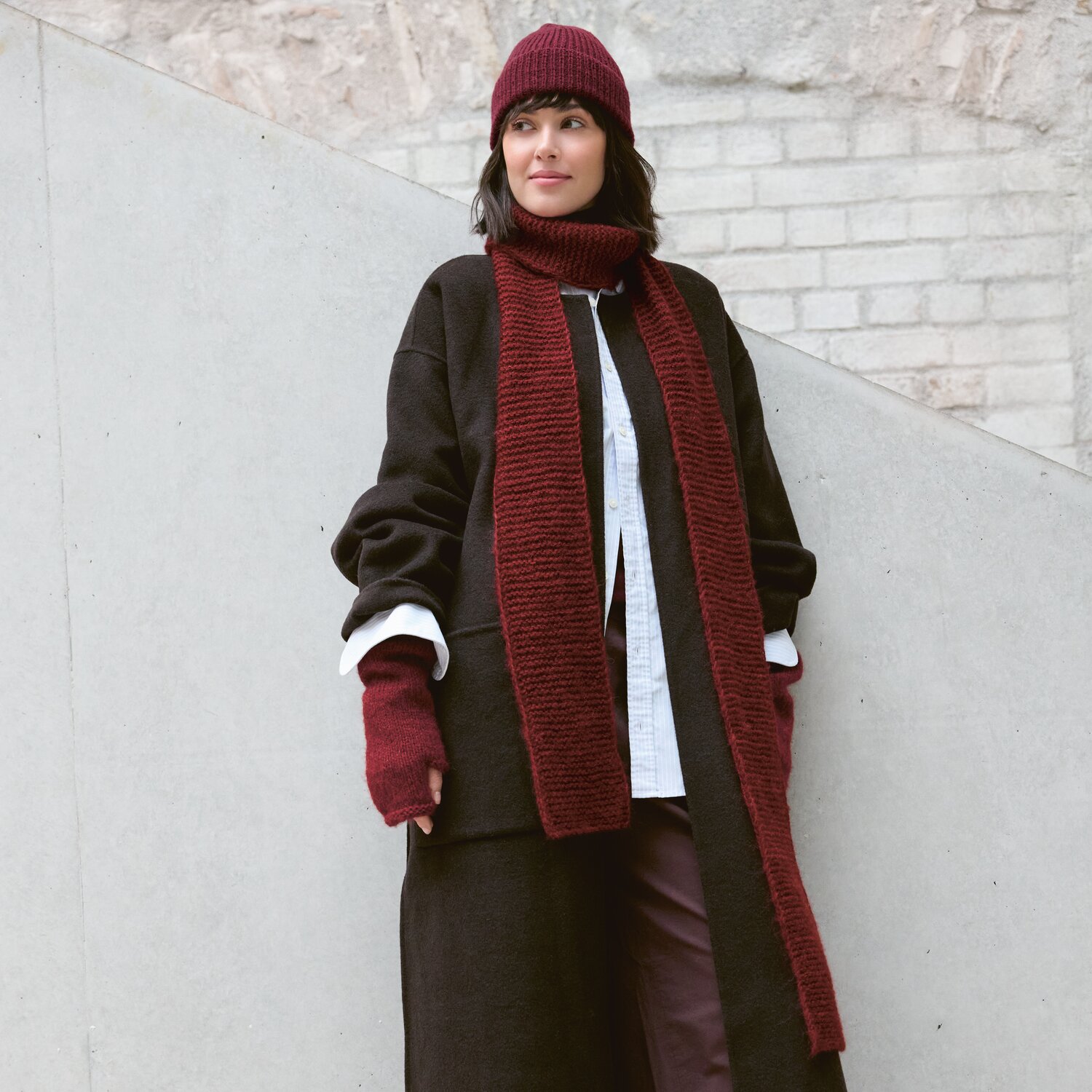 Strickset Schal Modell 17 aus Luxury Knits - Premium Natural Fibres von Rico Design Strickset Schal Modell 17 aus Luxury Knits: Frau trägt roten Schal, Mütze und Handschuhe, kombiniert mit dunklem Mantel vor heller Wand.