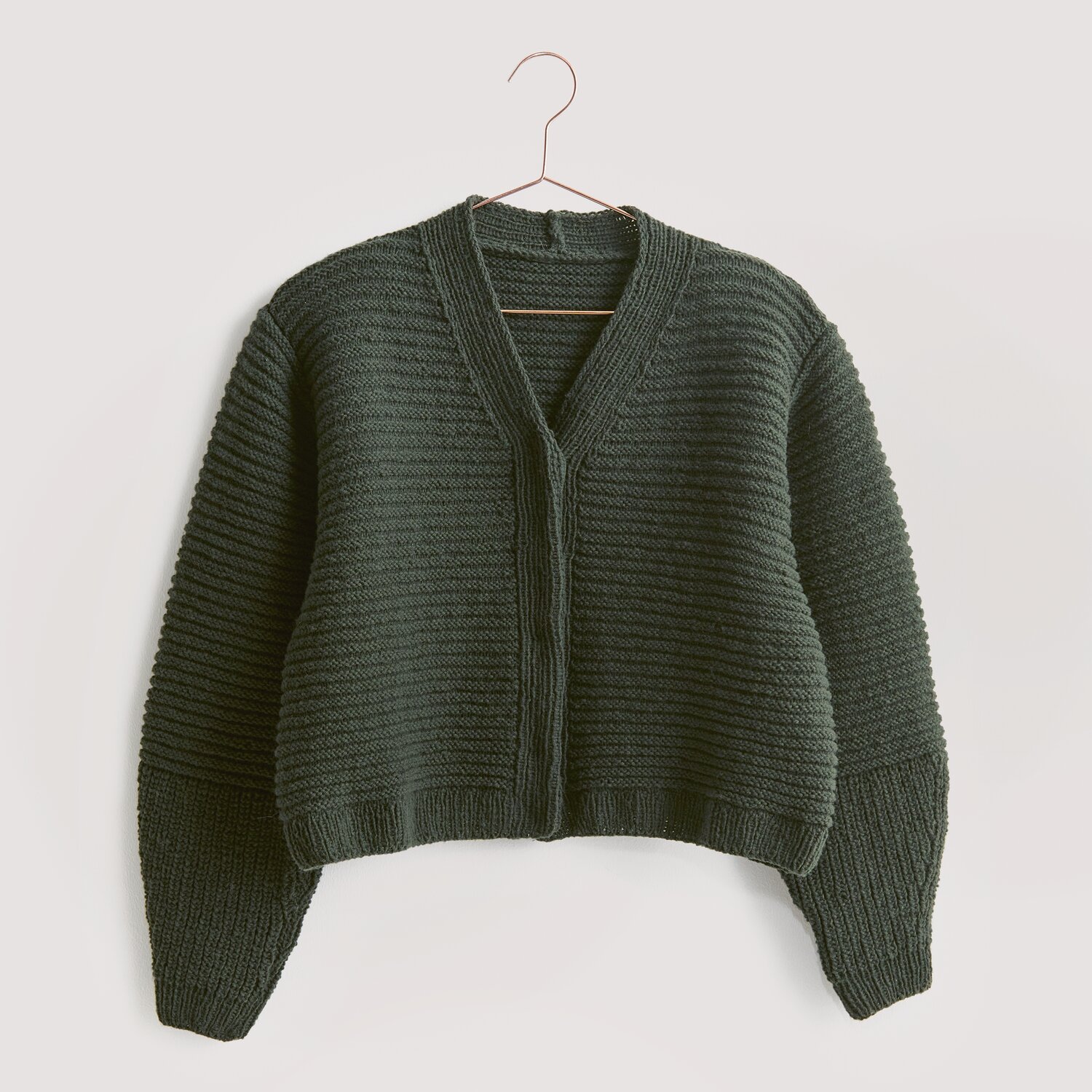 Luxury Knits - Premium Natural Fibres Sprachauswahl deutsch von Rico Design Strickset Jacke Modell 16 aus Luxury Knits, dunkelgrün, aus Premium Natural Fibres, auf einem Kleiderbügel.