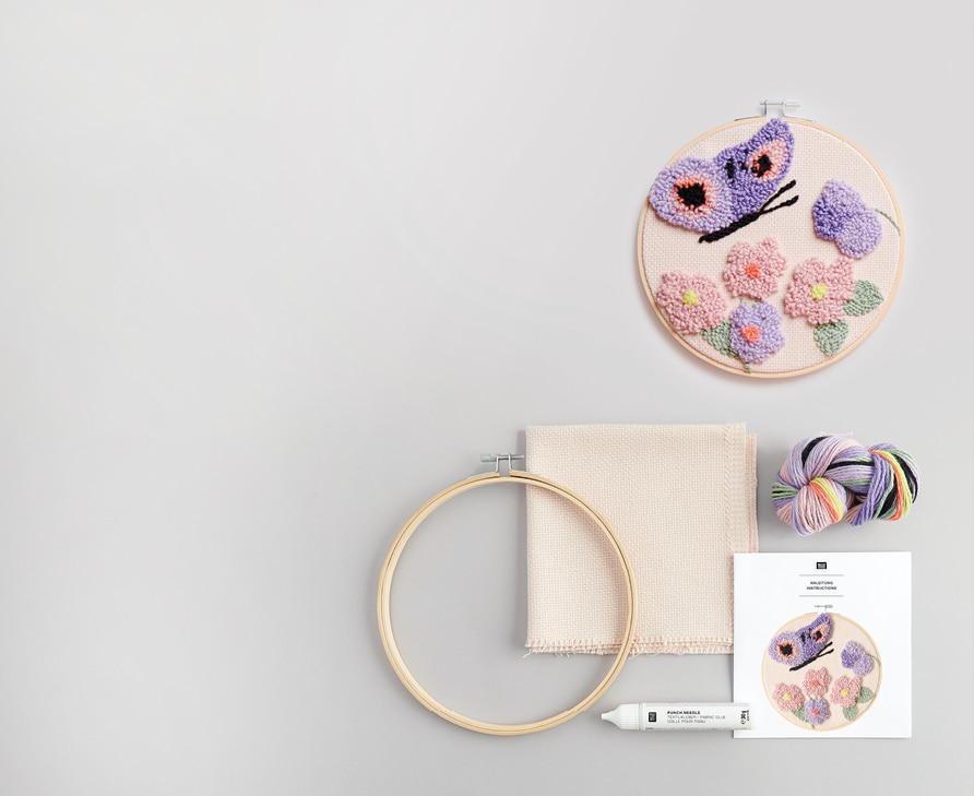 Punch Needle Bild mit Blumen und Schmetterling liegt neben Stickrahmen, Anleitungsbogen, Wolle und Stoff auf grauem Hintergrund angeordnet