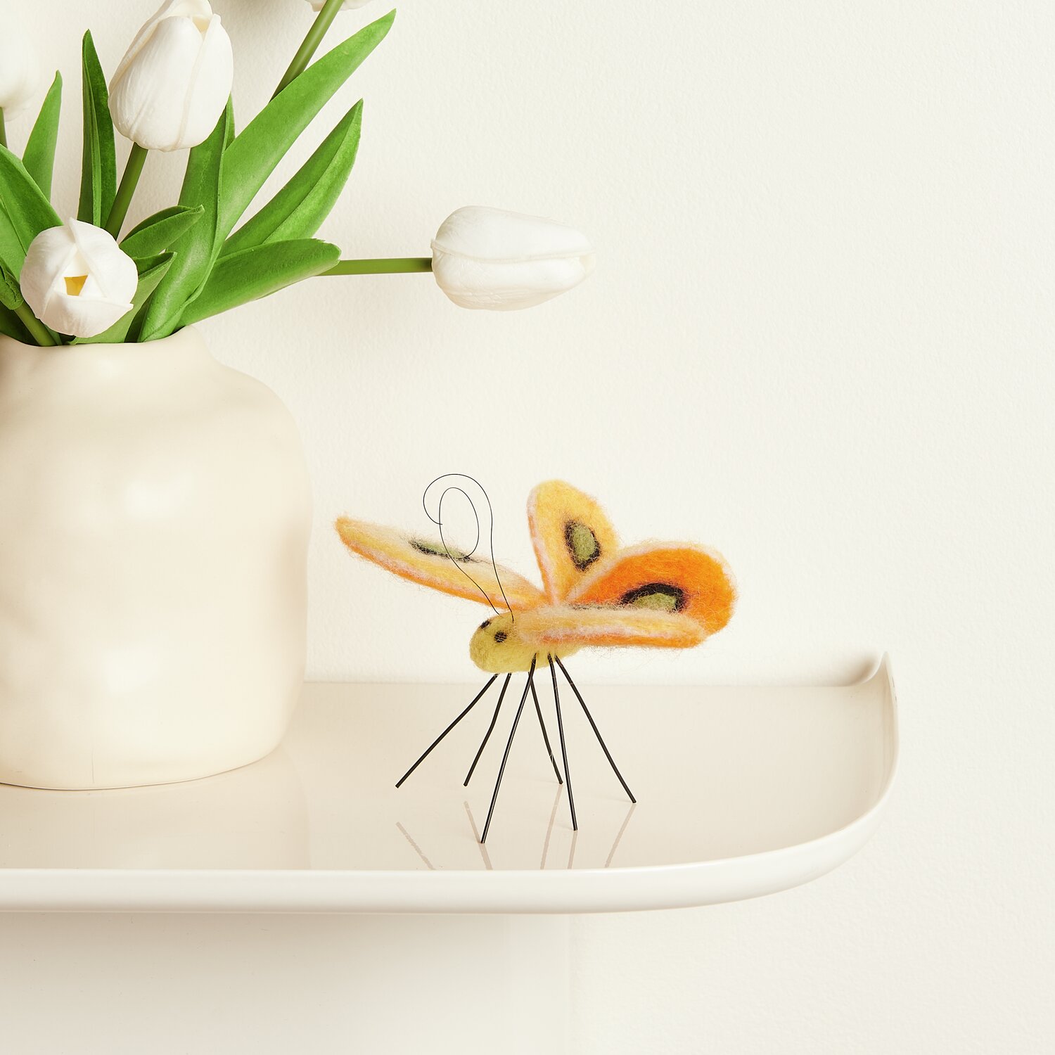 Aufhänger Filz-Schmetterling: Gelber Filz-Schmetterling von Rico Design auf einem Regal neben einer Vase mit Tulpen. Größe 10x10,5x7cm.