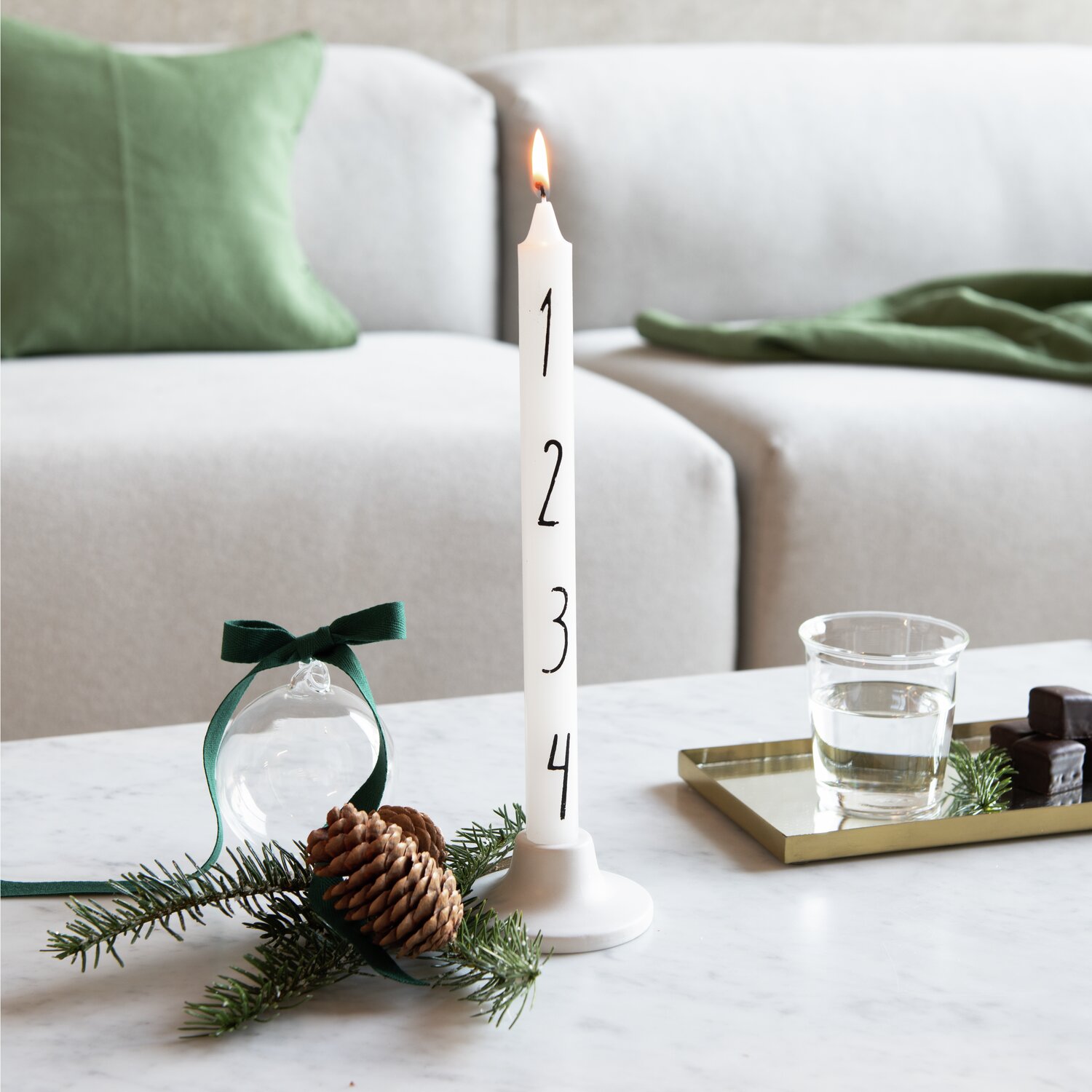 Anwendungsbild. Eine weiße Adventskerze von Rico Design mit den Zahlen 1 bis 4 steht auf einem Tisch. Im Hintergrund ist ein Sofa mit grünen Kissen zu sehen.