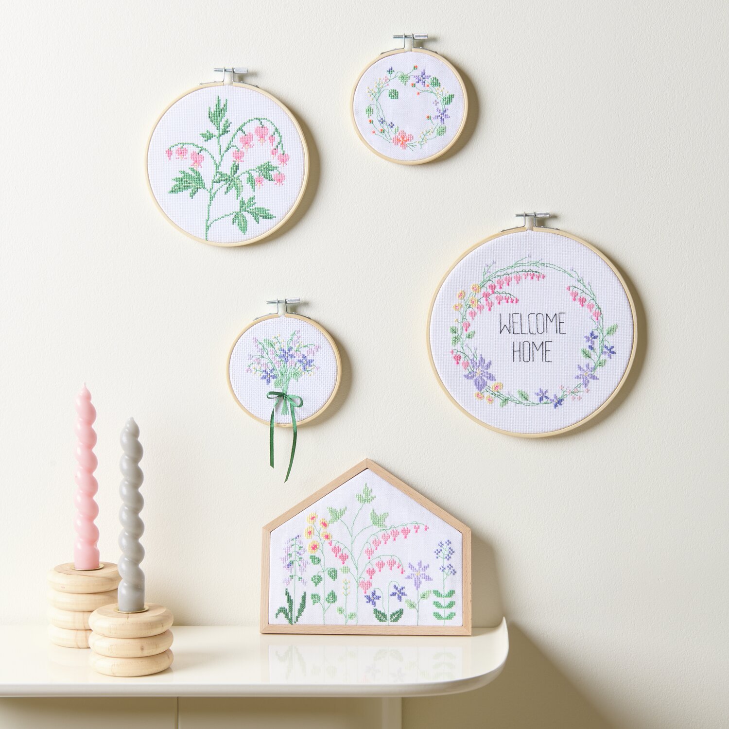 Stickpackung Gezählter Kreuzstich Blumenwiese: Vier Stickrahmen mit floralen Motiven und pastellfarbenen Kerzen auf einem Tisch.
