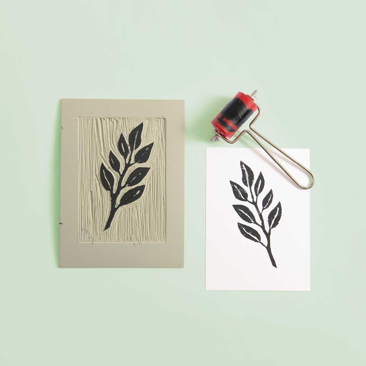 Eine Linolplatte mit Blättern eingeschnitzt, eine Linolwalze und ein bedrucktes Papier liegen auf grünem Hintergrund.