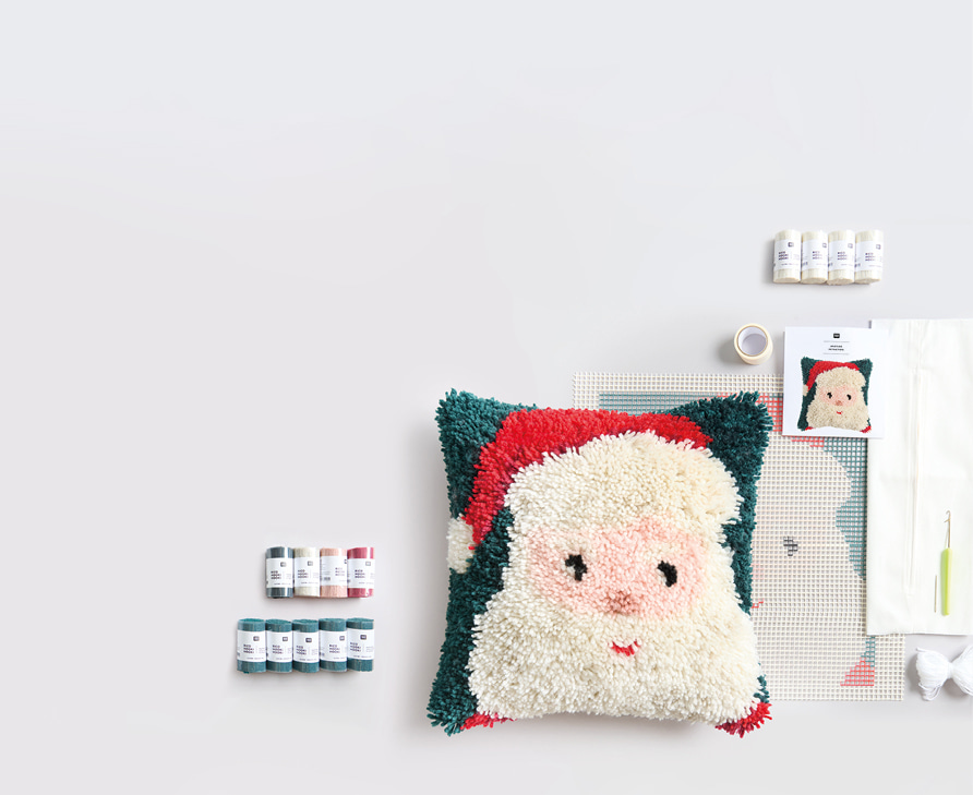 Ein geknüpftes Kissen mit Santamotiv, Knüpfgarn, Haken und weiteres Knüpfzubehör liegen ausgebreitet auf grauem Hintergrund. 
