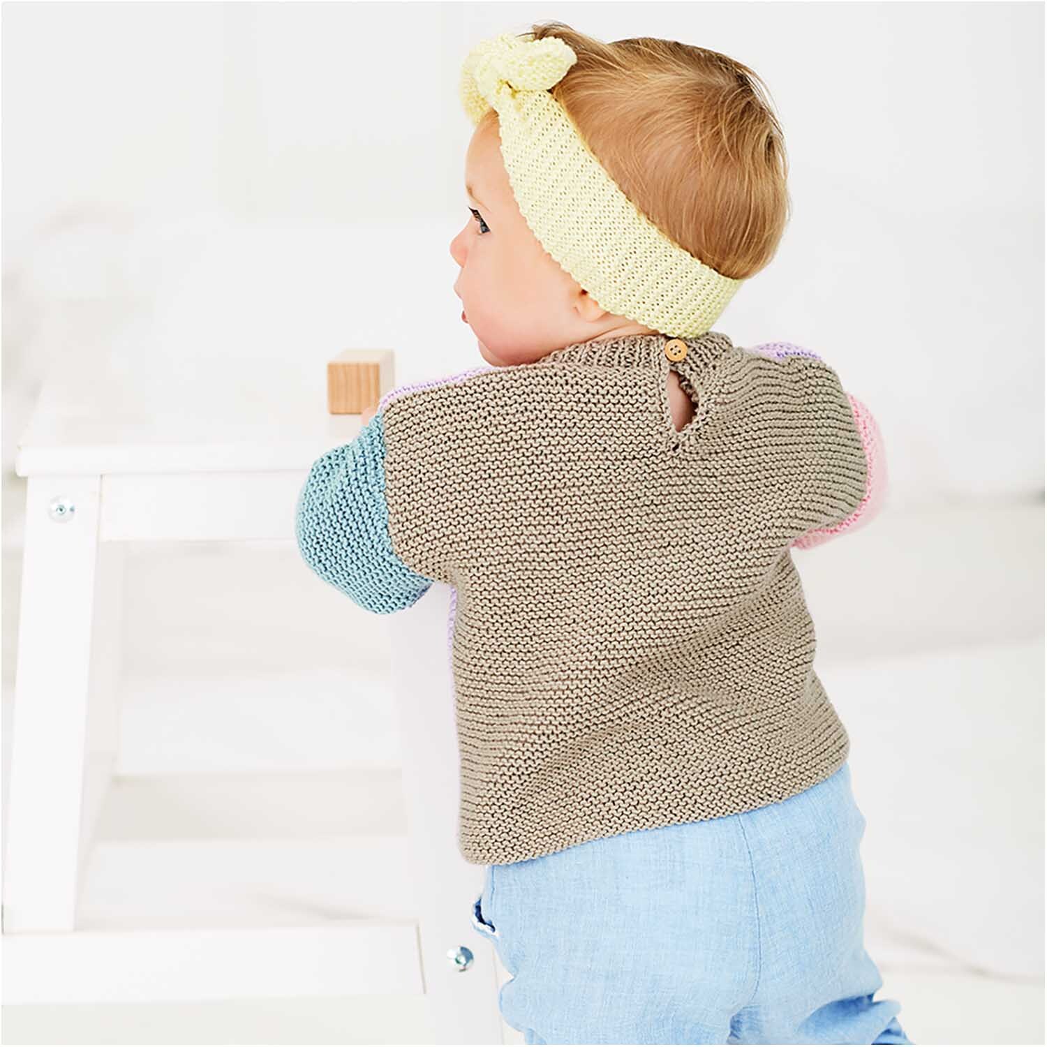 Rico Baby Cotton Soft dk: Ein Baby trägt einen gestrickten Pullover mit bunten Ärmeln und ein pastellgelbes Stirnband.