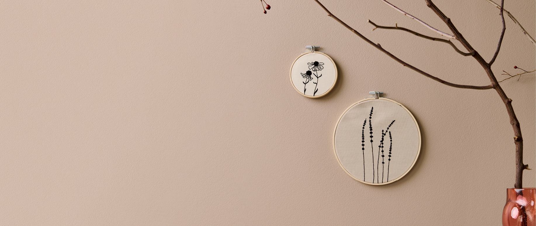 Stickbilder mit Gräser- und Blumenmotiven hängen an einer beigen Wand. 