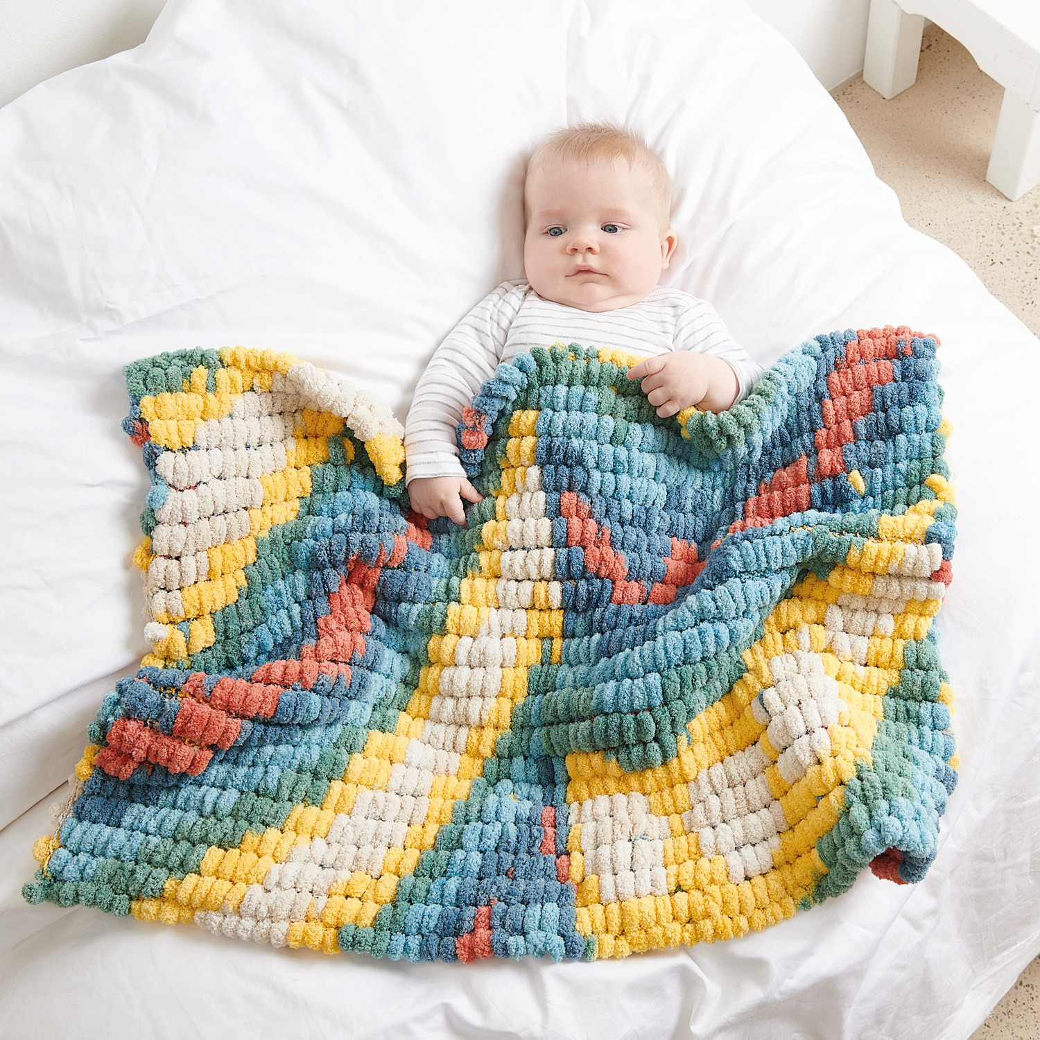 Creative Pompon Print: Baby liegt auf weißem Bett mit bunter, weicher Decke von Rico Design.