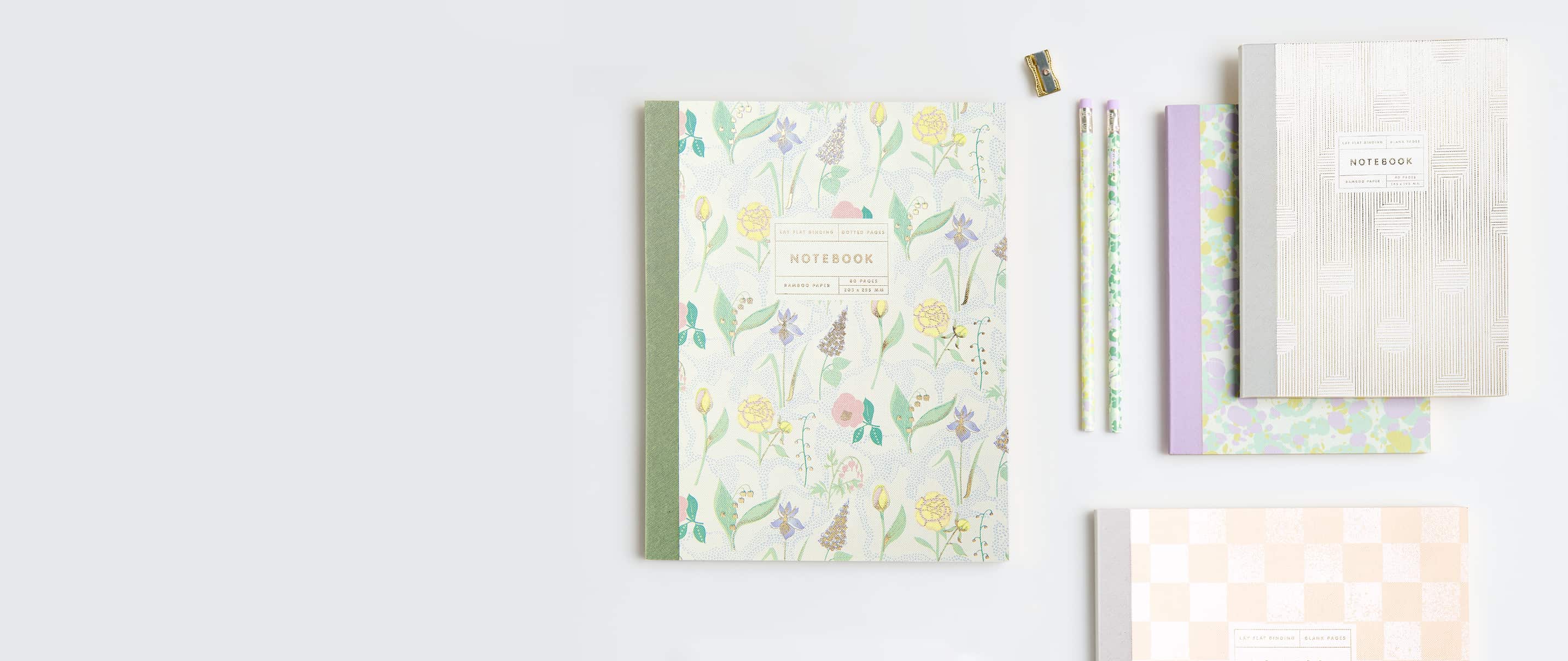 Notizbücher in Pastelltönen, floralen und abstrakten Mustern liegen umgeben von Bleistiften auf grauem Hintergrund.