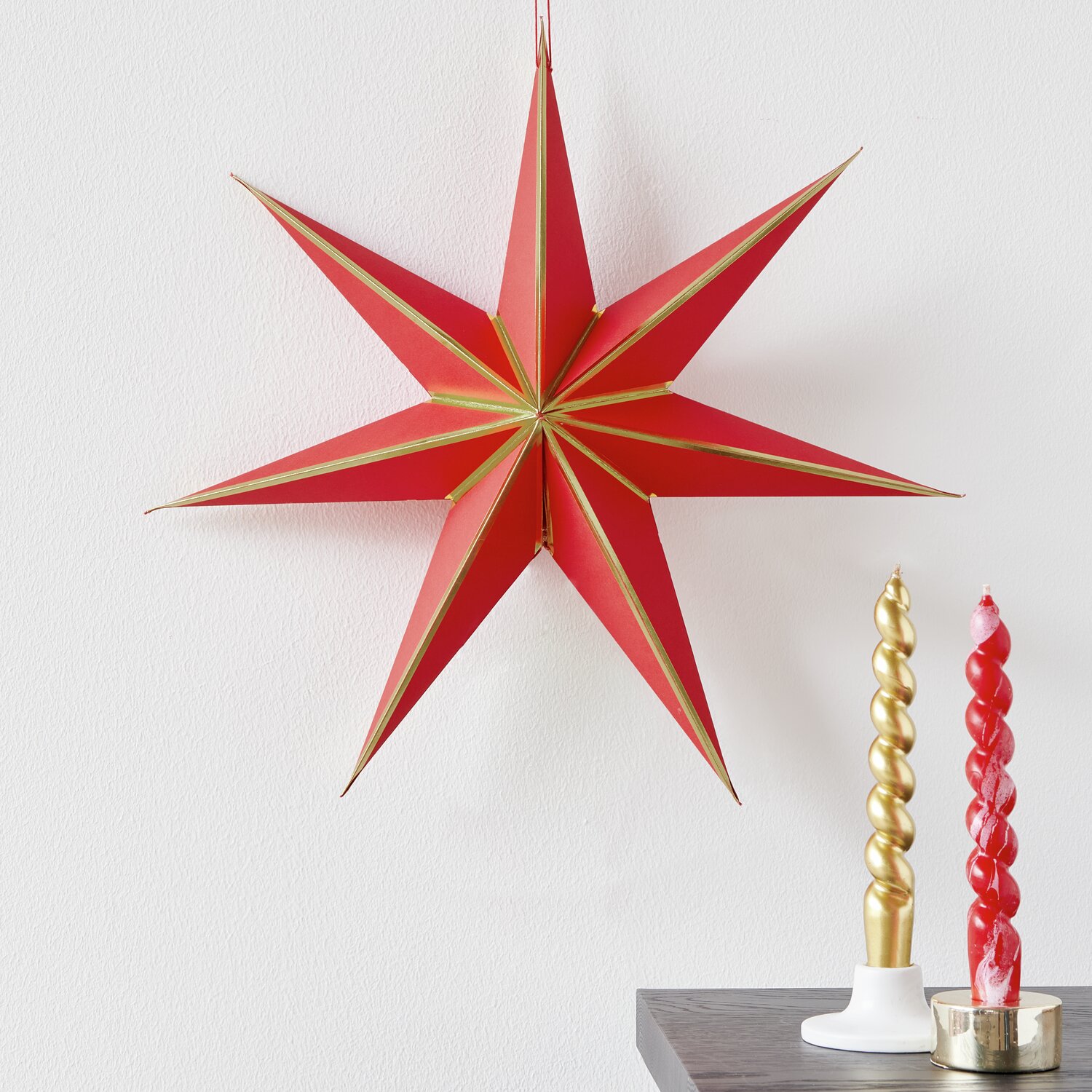 Papierstern in Rot/Gold von Rico Design, Ø45cm, dekoriert neben gedrehten Kerzen auf einem Tisch.