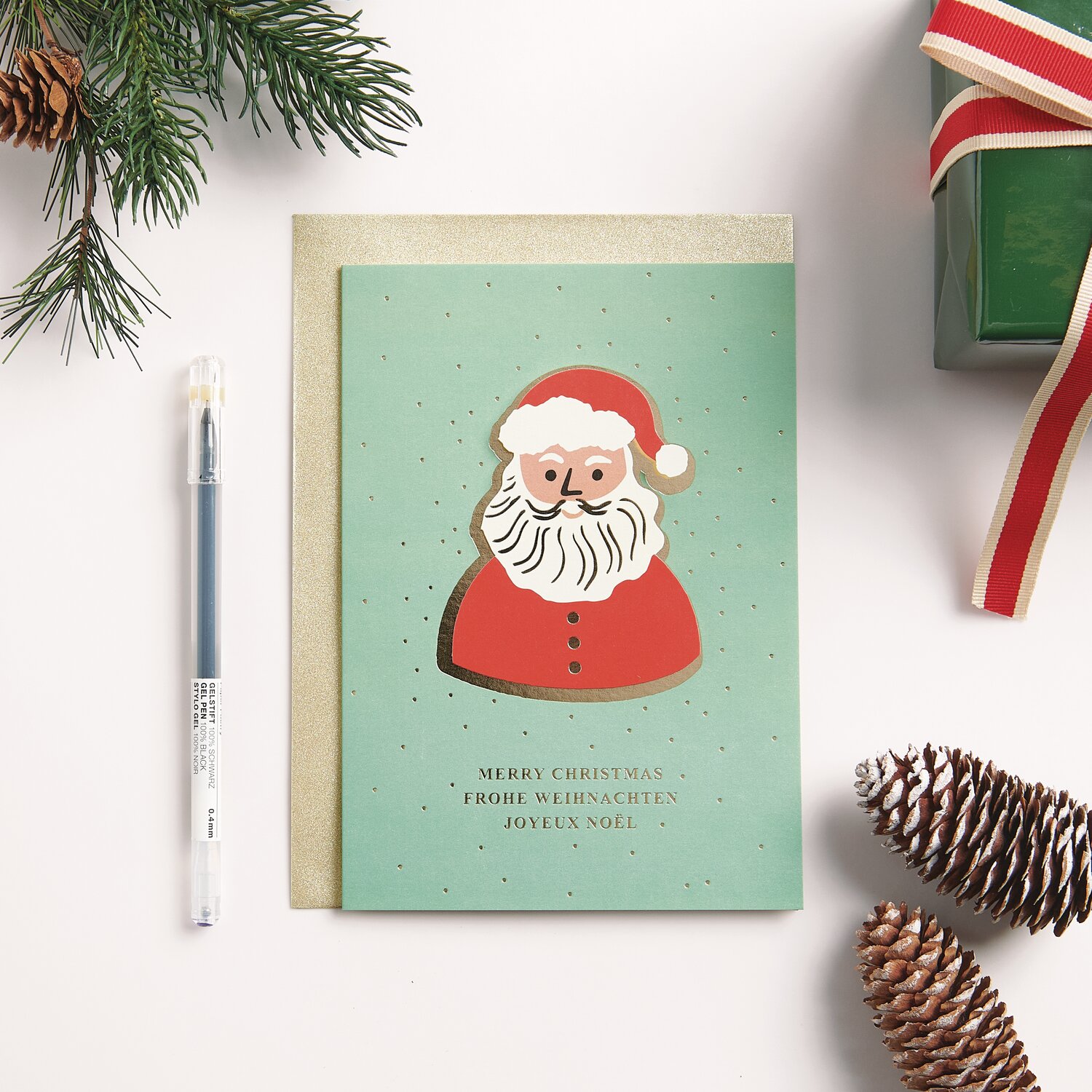 3D-Kartenset Weihnachtsmann B6 12-teilig von Rico Design 3D-Kartenset Weihnachtsmann B6: Weihnachtskarte mit rotem Santa-Motiv auf grünem Hintergrund, umgeben von Tannenzweigen und Geschenk.