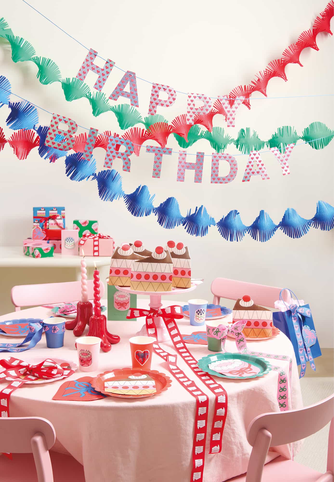 Ein gedeckter Tisch mit bunten Papptellern und Servietten in Tortenform. Darüber hängen bunte Kreppgirlanden und eine Happy-Birthday-Girlande.