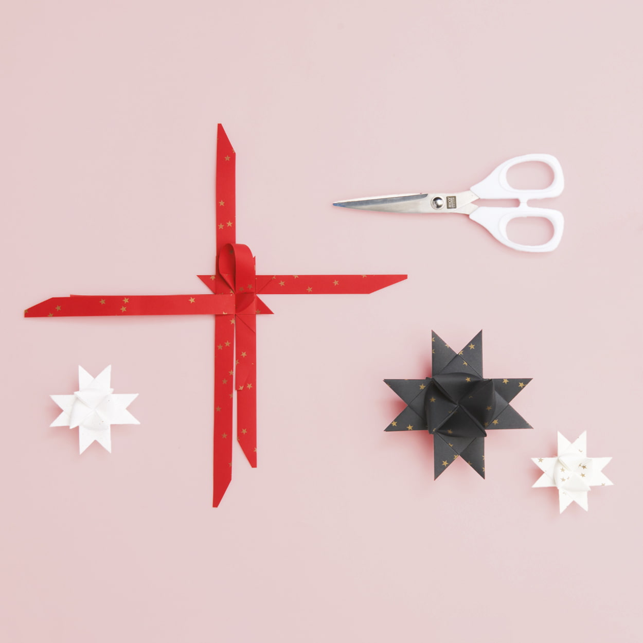 Gefaltete Fröbelsterne in Rot, Schwarz und Weiß und eine Schere liegen auf rosa Hintergrund.