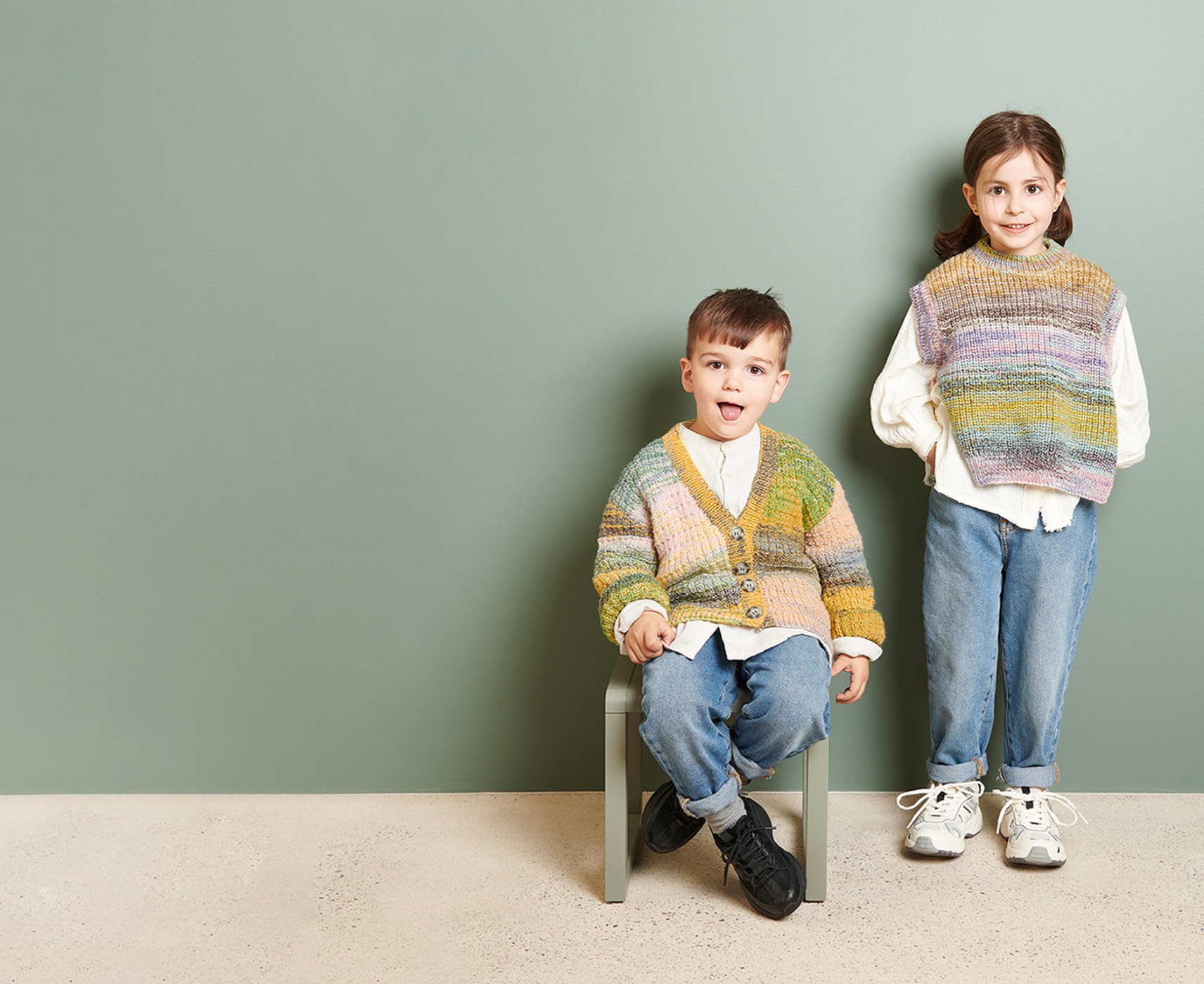 Drei Kinder in gestrickten Jacken und Pullundern des Strickhefts Rico Kids 13 gekleidet, posieren vor einer dunkelgrünen Wand.