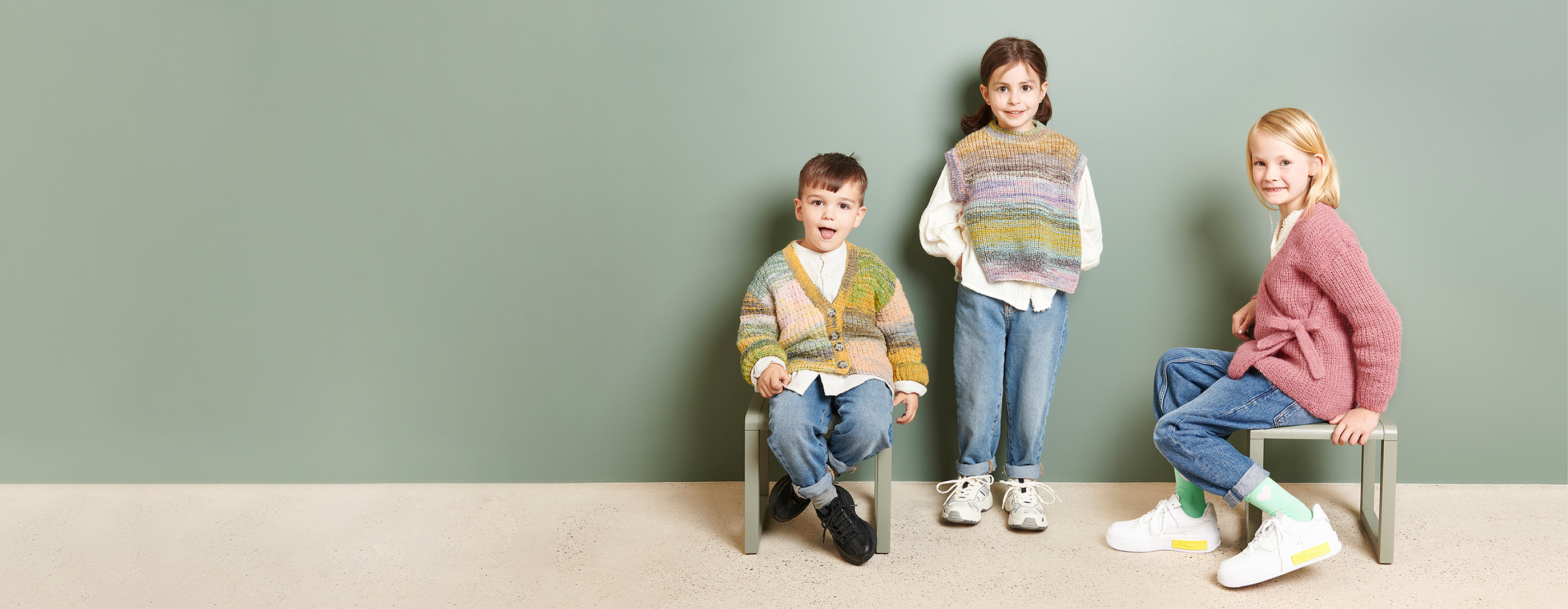 Drei Kinder in gestrickten Jacken und Pullundern des Strickhefts Rico Kids 13 gekleidet, posieren vor einer dunkelgrünen Wand.