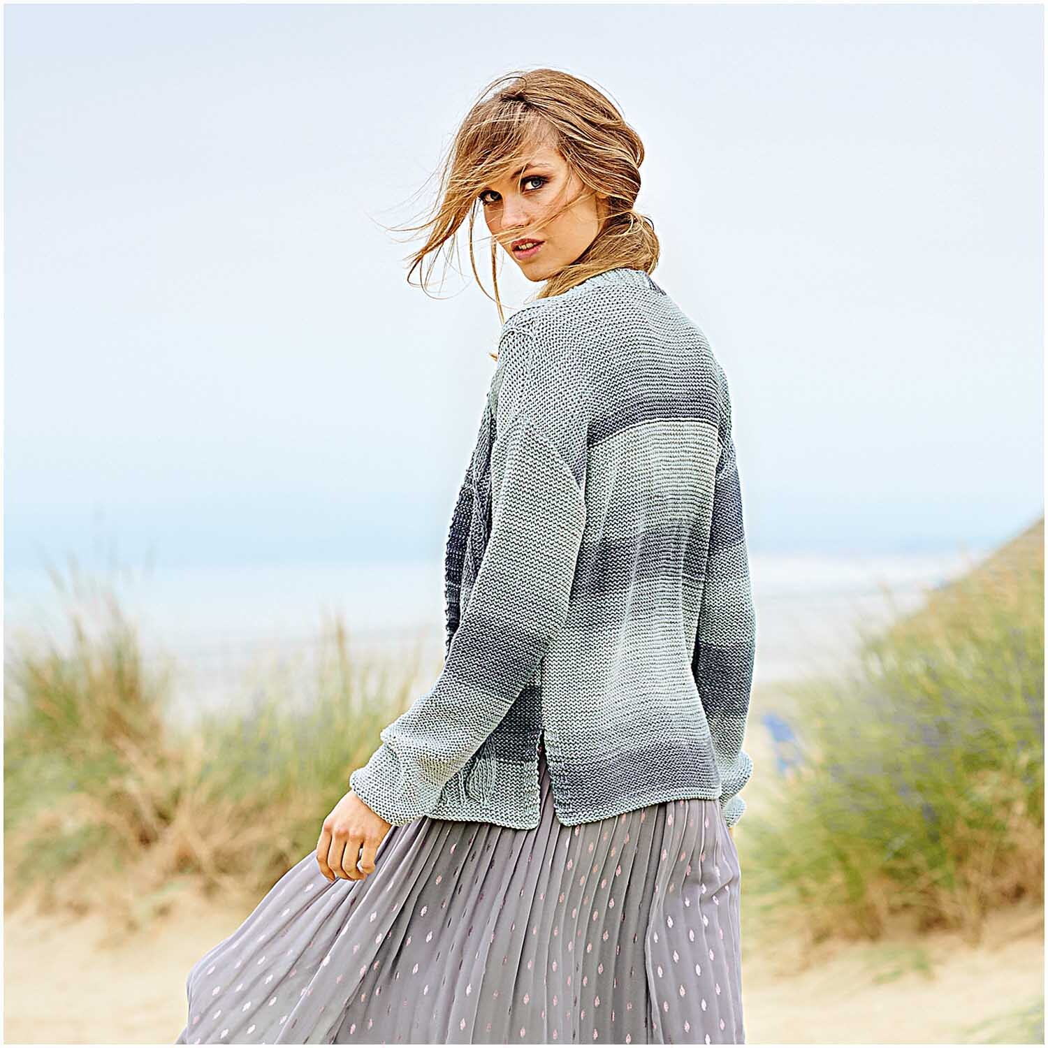 Fashion Colour Spin: Frau in gestricktem Cardigan von Rico Design am Strand, mit wehendem Haar.