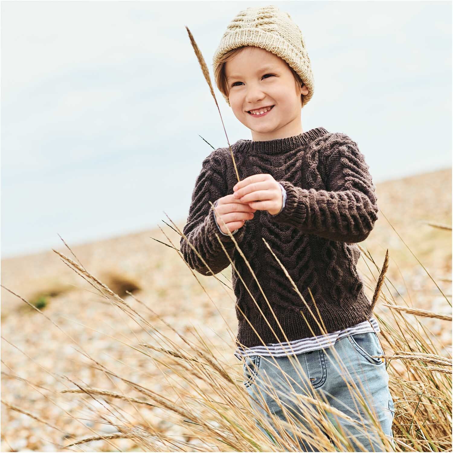 Essentials Merino dk: Kind in gestricktem Pullover am Strand, lächelnd, mit Grashalm in der Hand.