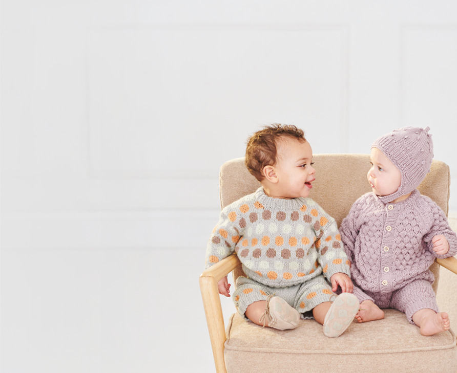 Zwei Babys sitzen in Strickkleidung gekleidet auf einem Stuhl und schauen sich fröhlich an