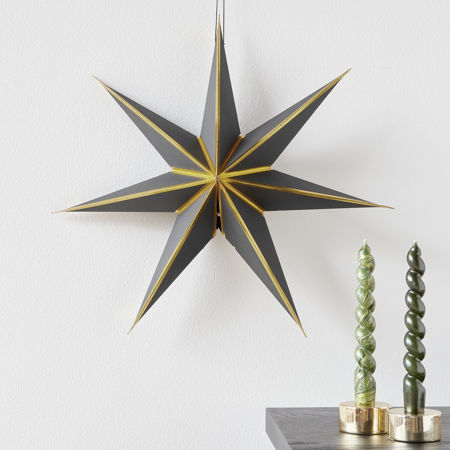Papierstern, Ø45cm, in Schwarz/Gold von Rico Design, hängt dekorativ neben grünen Kerzen.