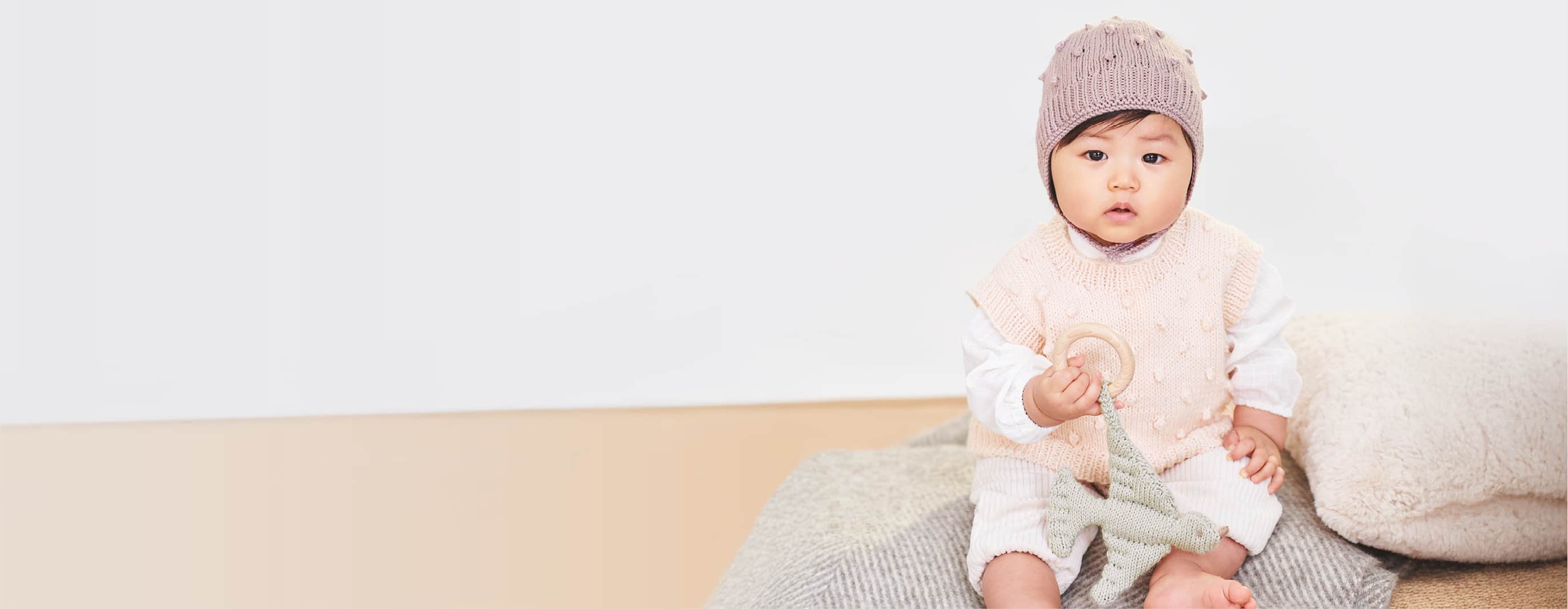 Ein Baby sitzt mit gestricktem Pullover und gestrickter Mütze auf einer Decke und hält einen Beißring in der Hand