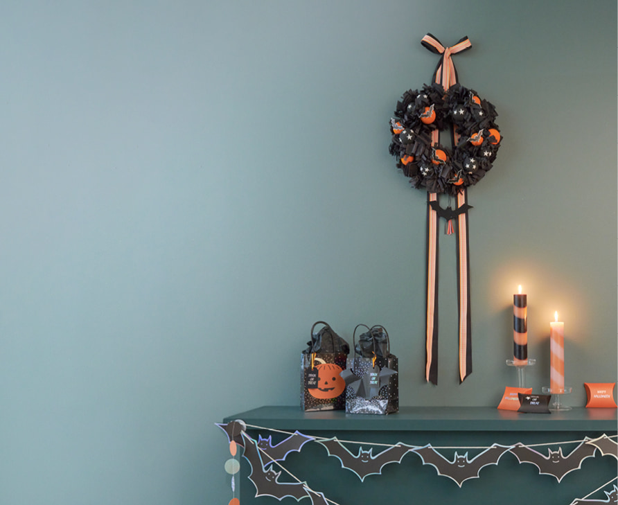Ein Kaminsims ist mit Halloween-Artikeln dekoriert. Geschenktüten, Kerzen, Girlanden und ein Kranz in Schwarz und Orange schmücken die Szene