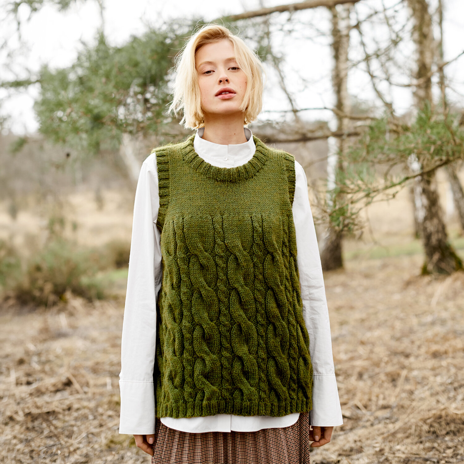 Strickanleitung Pullunder aus Soft Alpaca, moderner Zopfmuster-Slipover in Moosgrün, getragen von Frau über weißer Bluse in herbstlicher Waldlandschaft
