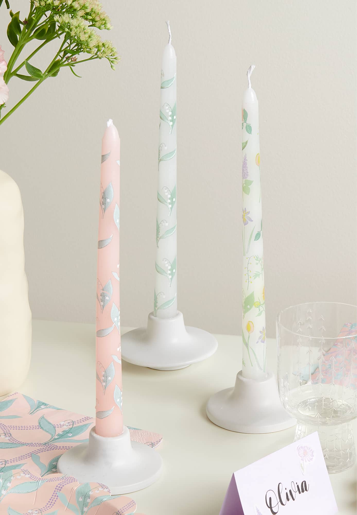 Drei Stabkerzen in Pastelltönen mit Blumenmotiven stehen in Kerzenhaltern dekoriert auf einem Tisch.