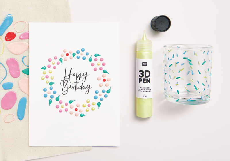 Bemalte Happy Birthday-Karte mit 3D Punkten liegt zusammen mit einem 3D Pen auf grauem Hintergrund.