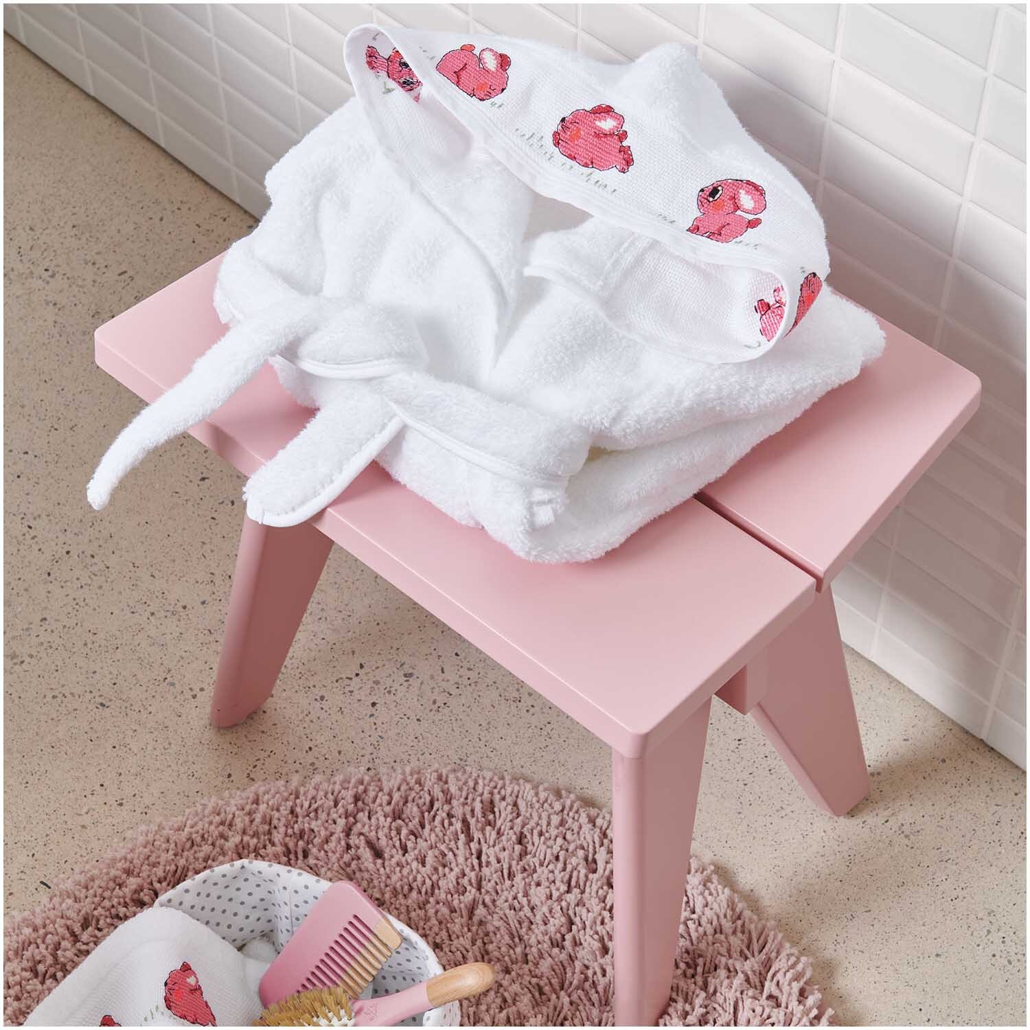 Kinderbademantel ab 18 Monate Farbe Weiß von Rico Design Kinderbademantel ab 18 Monate auf rosa Hocker, mit Kapuze und niedlichen roten Motiven. Ideal für Kleinkinder.