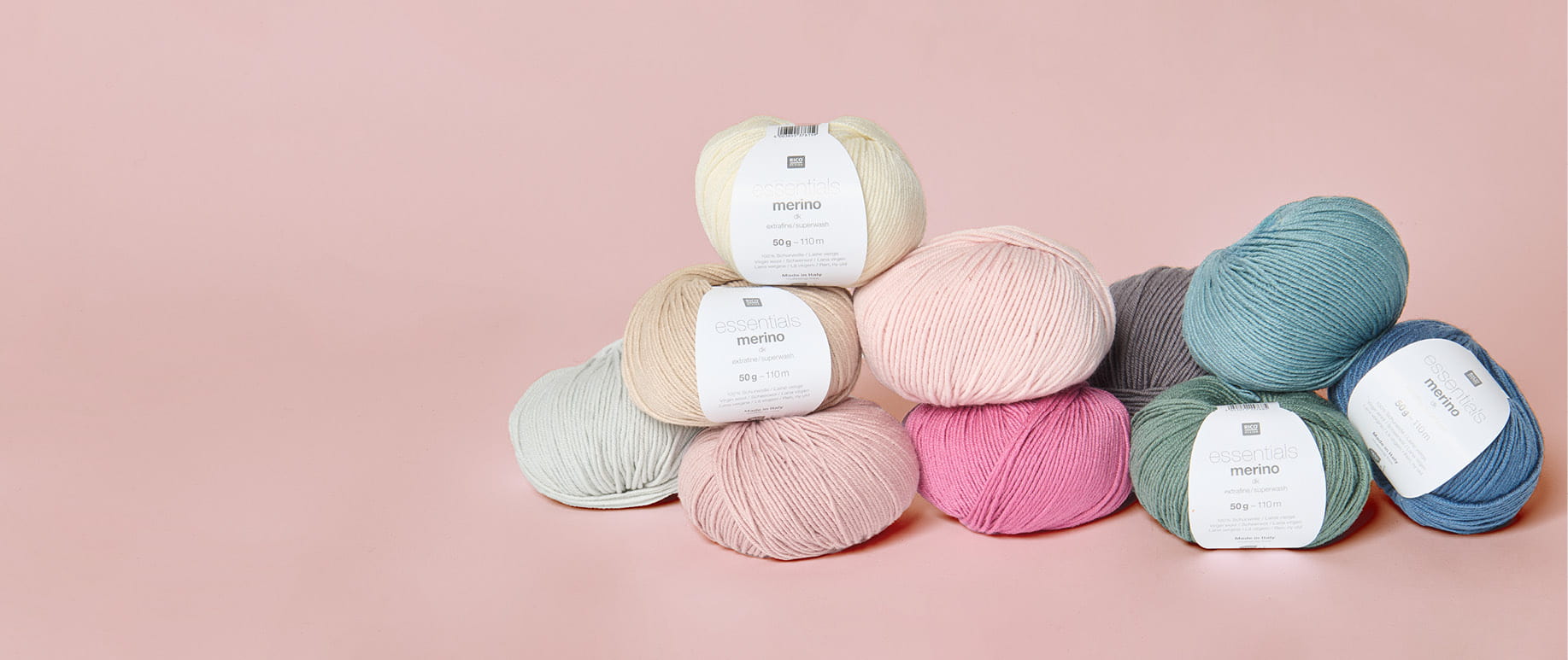 Mehrere Knäuel von Essentials Merino dk in verschiedenen Farben liegen gestapelt vor rosa Hintergrund. 