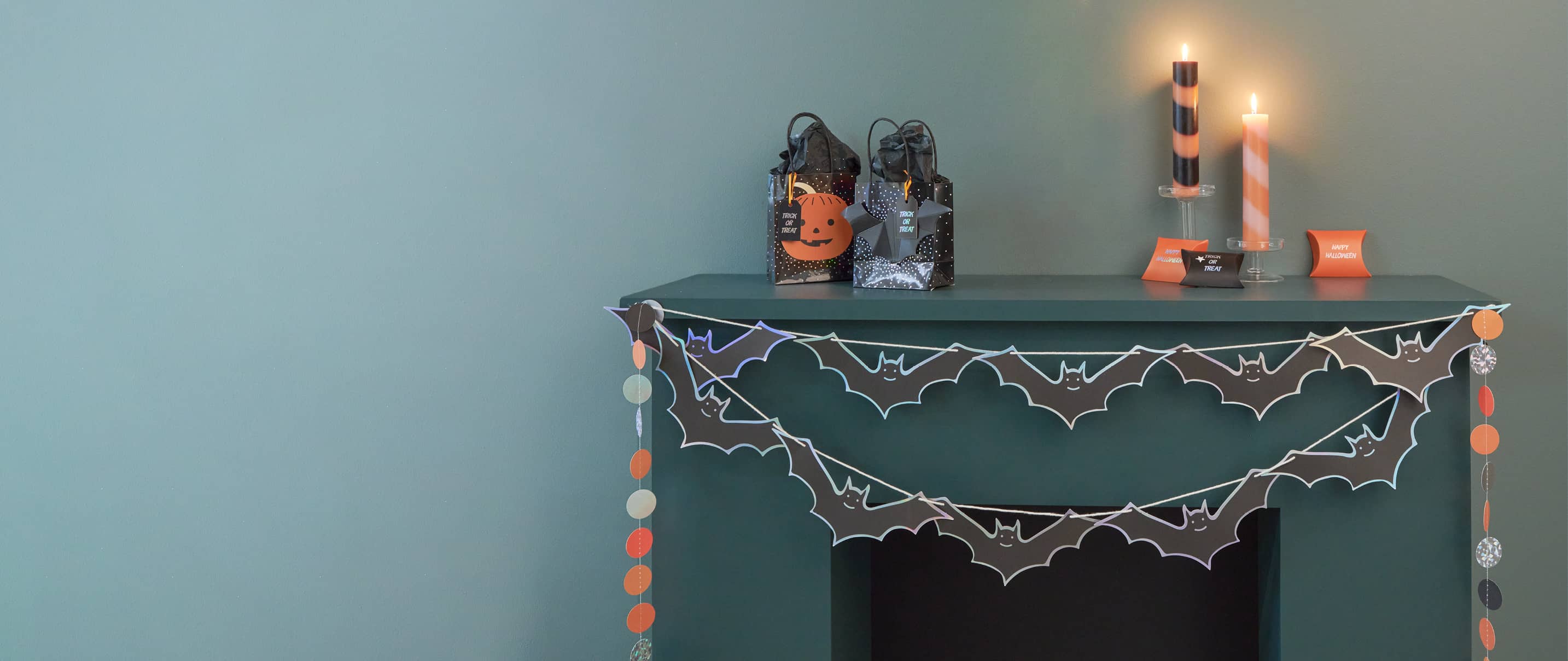 Ein Kaminsims ist mit Halloween-Artikeln dekoriert. Geschenktüten, Kerzen und Girlanden in Schwarz und Orange schmücken die Szene