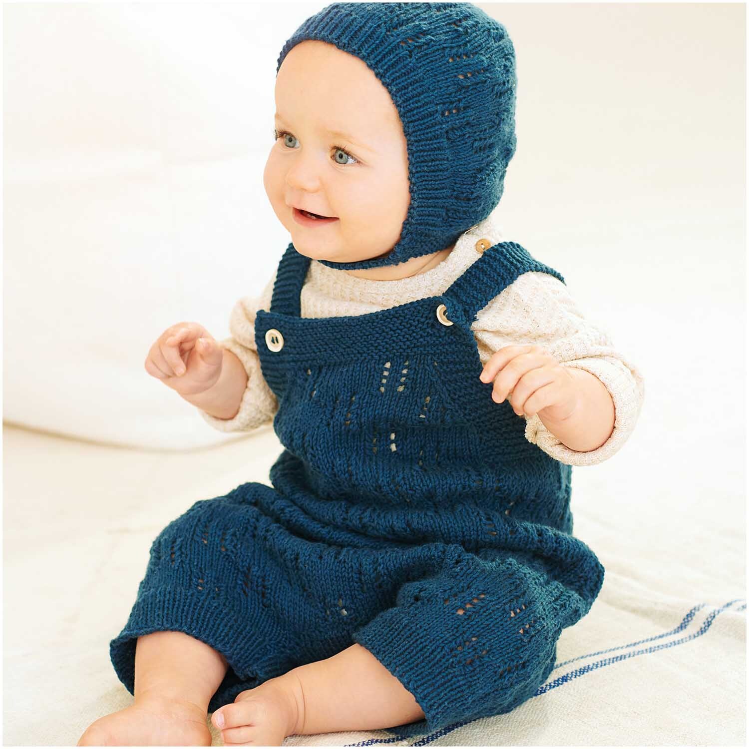 Baby in gestricktem marineblauem Outfit aus Bio-Baumwolle von Rico Design, sitzt lächelnd auf hellem Hintergrund.