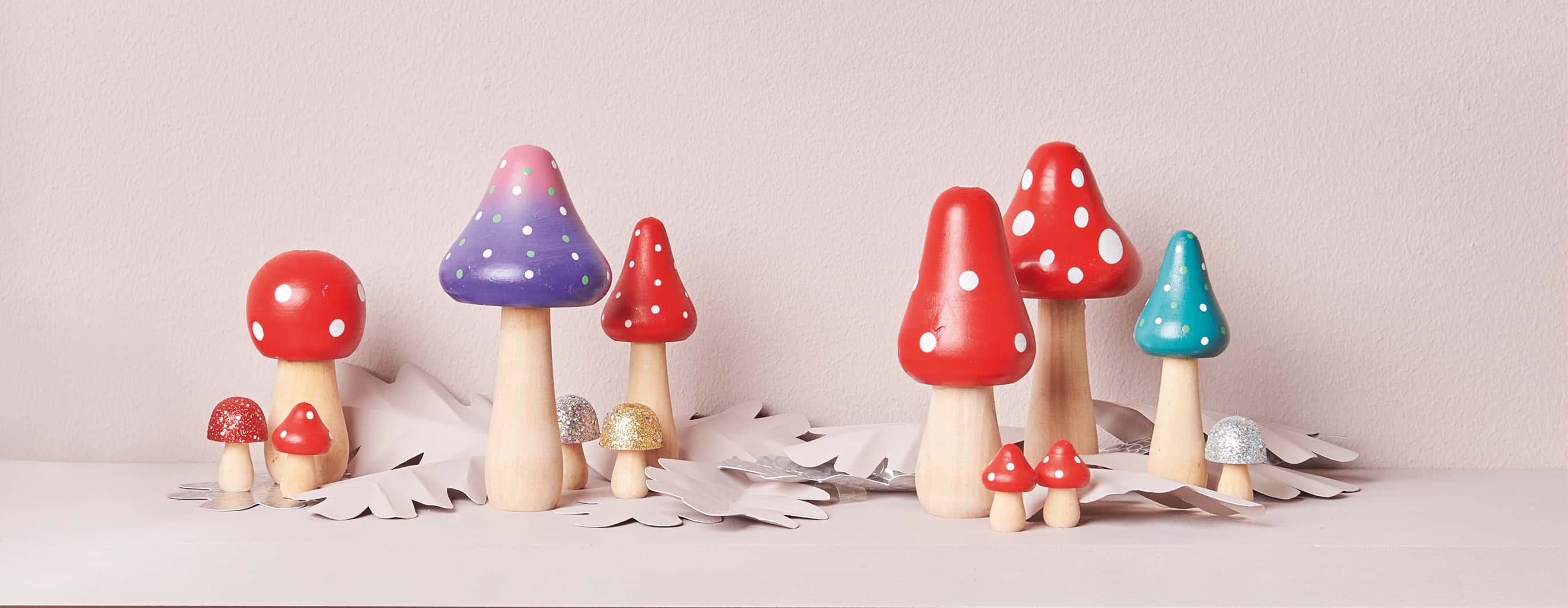 Fliegenpilze aus Holz in verschiedenen Farben, Formen und Größen stehen herbstlich dekoriert vor rosa Hintergrund