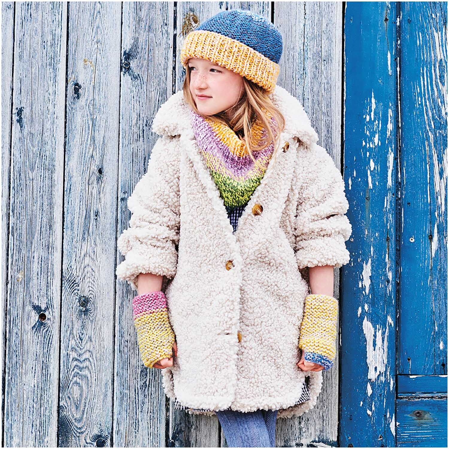Creative Chic-Unique: Mädchen in bunter Strickmütze und Schal, vor Holzwand stehend, trägt flauschigen Mantel.