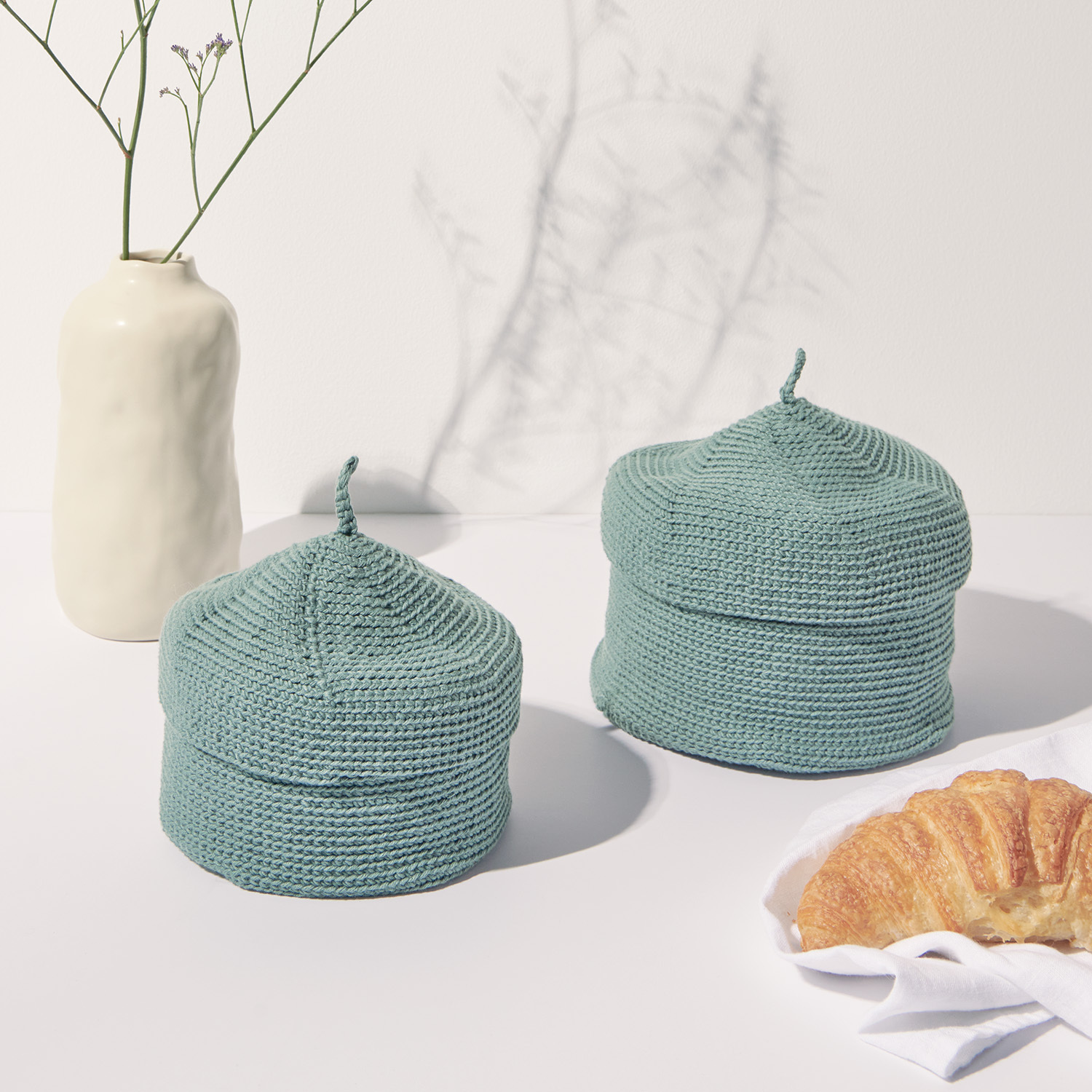 Häkelanleitung Korb mit Deckel aus Essentials Organic Cotton aran: Zwei gehäkelte Körbe in sanftem Grün neben einer Vase und Croissants auf einem Tisch.